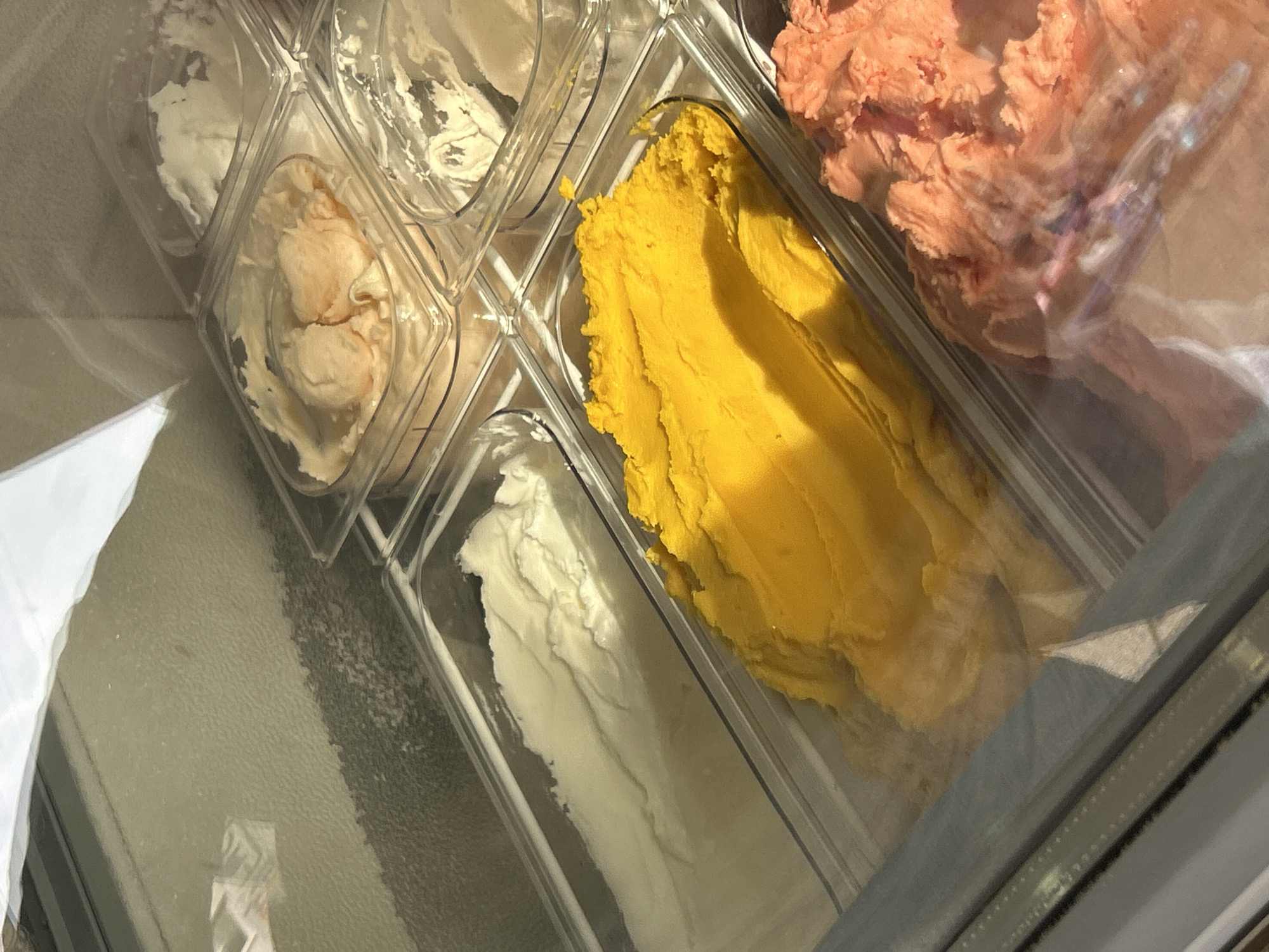 The image shows a display of various flavors of ice cream in clear plastic tubs, likely in a frozen dessert shop. The tubs are arranged in a refrigerated display case. In the foreground, to the left, is a tub of white ice cream. To its right and slightly behind is a tub filled with bright yellow ice cream, with visible texture suggesting it has been scooped. Further to the right, and partially visible in the foreground, is a tub of pink ice cream with a rough texture. In the background, several other tubs of ice cream are visible, including one with a peachy-orange color and another with white ice cream. The scene is illuminated by overhead lighting, causing reflections on the glass of the display case and the ice cream itself. The overall impression is that of a selection of cold, creamy desserts. The location is stated to be Waldbröl, Germany.