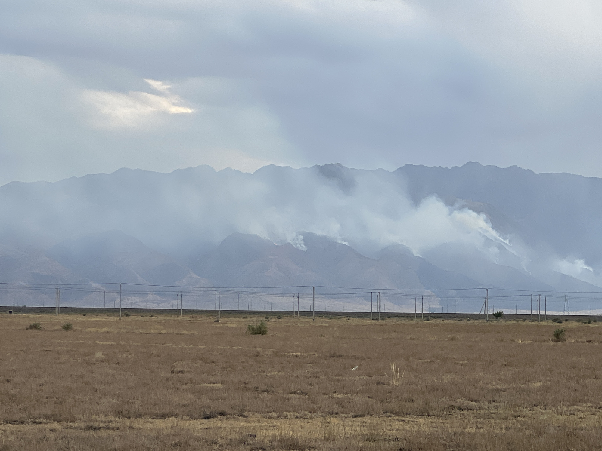 The image shows a wide, dry, grassy field in the foreground. In the middle ground, there is a line of utility poles with wires stretching across the landscape, and a darker strip of land behind them. The background features a mountain range partially obscured by smoke, indicating a fire. The sky is overcast with some patches of brighter light breaking through the clouds. The scene appears to be an outdoor, rural or semi-rural environment.