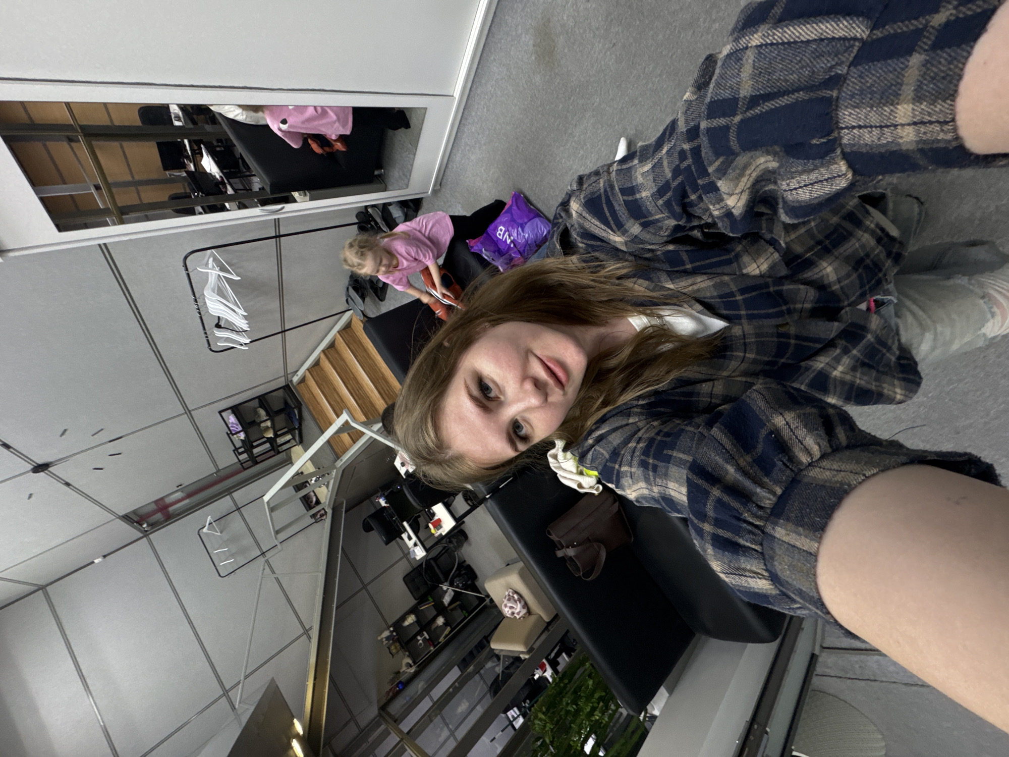 The image is a selfie of a young woman wearing a blue and beige flannel shirt. She is seated on a black chair. Behind her is a child in a pink shirt sitting on a similar black chair, interacting with an item on the chair. Adjacent to the child is a purple shopping bag. The setting appears to be a dressing room or backstage area, with clothing racks and mirrors visible. In the background, there is a staircase and other chairs, suggesting a commercial or public space. The location is Perm, Russia.