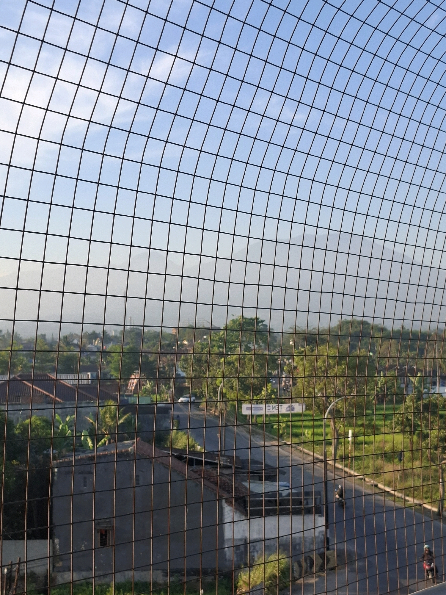 The image depicts a view of a suburban area in Sukabumi, Indonesia, seen through a wire mesh fence. In the foreground, the fence creates a grid-like obstruction. Beyond the fence, a residential area with houses, some with red tile roofs, is visible. Lush green trees and vegetation are scattered throughout the landscape. A road runs through the scene, with a few vehicles, including motorcycles, on it. In the background, mountains shrouded in a light haze are discernible against a clear blue sky with some wispy clouds. The overall impression is a peaceful, somewhat hazy morning or afternoon view of a town nestled amongst greenery and mountains.