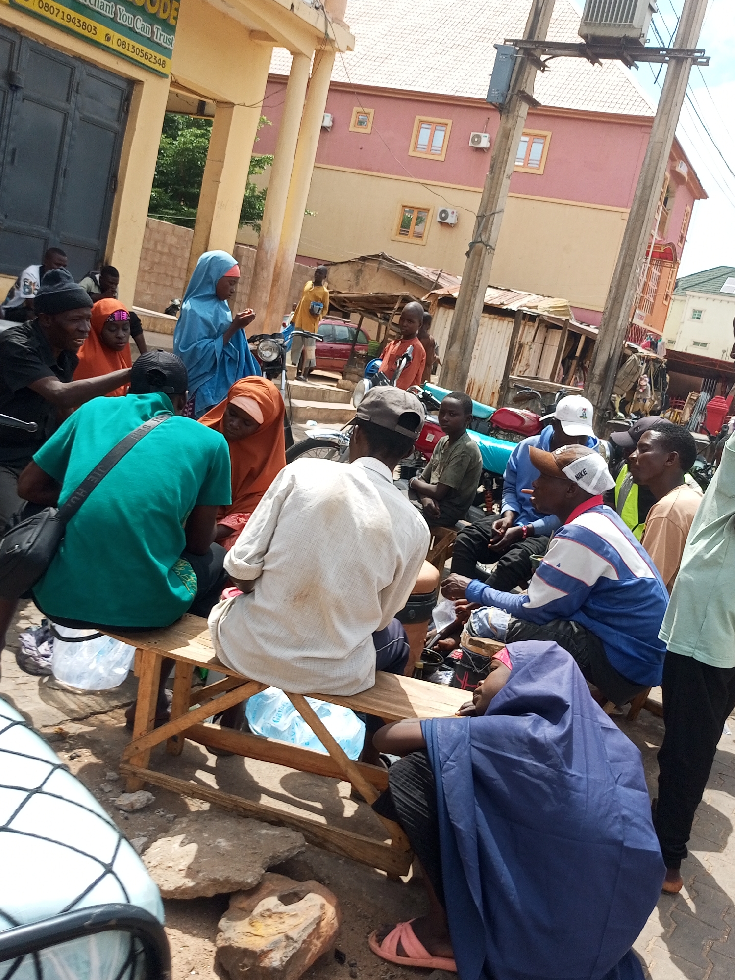 In the city of Arawa, Nigeria, a group of people are gathered outdoors in what appears to be a market or commercial area. The setting features a building with a yellow facade and pillars, suggesting a storefront or office. Behind the people, rudimentary structures and a multi-story building with pink and yellow paint are visible. Electrical poles and wires are also present, indicating urban infrastructure. The ground is paved with dirt and scattered rocks.
Several individuals are seated on a wooden bench. One person in a green shirt and black shoulder bag is facing away from the camera, talking to another person wearing an orange headscarf and headscarf. To the right, a person in a light-colored shirt is also seated. A person wearing a blue headscarf is standing, looking down.
Other individuals are standing or partially visible. A person in a blue jacket with white and blue stripes is seated, smoking a cigarette. To their right, a person in a tan shirt and a person in a light green shirt are present. A young boy in an orange shirt stands in the background. The presence of motorcycles suggests a mode of transportation common in the area. Water bottles are visible on the ground and under the bench. The overall scene depicts a casual gathering in a busy, outdoor environment.