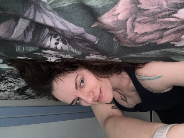 An indoor scene, identified as being in Boreti, Montenegro, features a single female subject lying on her left side on a bed. She possesses long, dark hair, and her face, presenting a slight smile, is directed towards the camera. The subject wears a dark blue tank top. Visible body markings include a black, stylized feather tattoo on the upper chest and a multi-colored striped tattoo (blue, orange, red) on the upper left arm. A white wristband is present on her raised right arm, suggesting camera operation. The bed is covered with a dark floral patterned sheet exhibiting green, purple, pink, and white motifs. A light blue horizontal panel is observable on the wall directly above the bed, against a light-colored background.