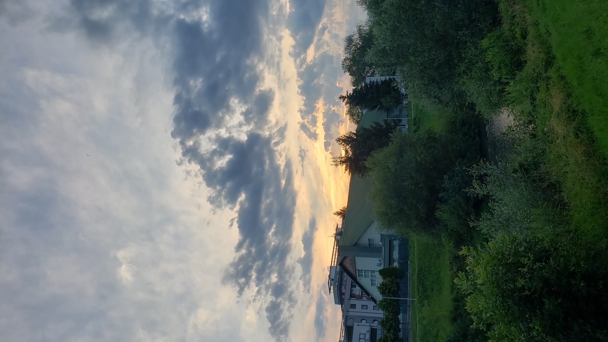 The image depicts a view of the city of Prešov, Slovakia, at dusk. The sky is filled with dramatic clouds, with the setting sun casting a warm, orange glow through a break in the clouds, illuminating the upper parts of some buildings and the sky. In the foreground, lush green trees and foliage line the banks of a body of water, likely the Torysa River, which meanders through the scene. Beyond the trees, several modern buildings are visible, including a multi-story residential building with a light-colored facade and a lower, greenish building with a prominent angled roof, possibly a public or commercial structure. The overall impression is one of a tranquil evening in an urban environment.