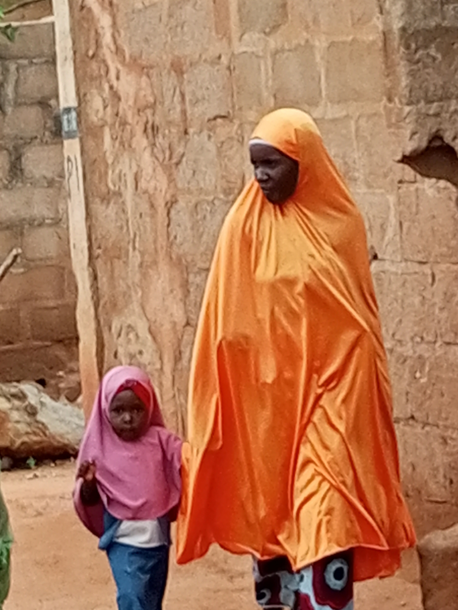 In Gombe, Nigeria, a young child wearing a pink hijab and blue trousers stands to the left of a woman in an orange hijab, looking towards the camera. The woman's face is partially obscured by her hijab, but she appears to be looking to the side. They are standing outdoors in front of a rough, brick wall.