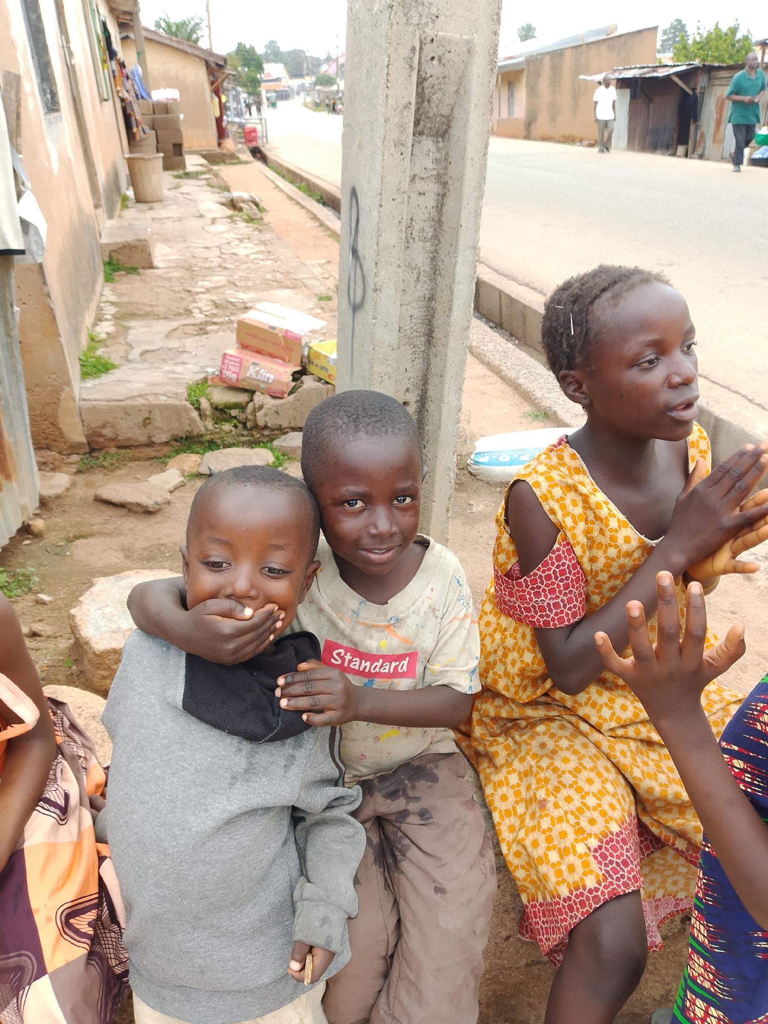 Three children are sitting on a low wall next to a street in what appears to be a city. The child on the left, wearing a gray hooded sweatshirt, has their hand over their mouth. The child in the middle, wearing a light-colored t-shirt with red writing, is leaning on the child on the left and appears to be looking at the camera. The child on the right, wearing a yellow and orange patterned dress, is clapping their hands. A street with some traffic and pedestrians can be seen in the background, along with buildings.