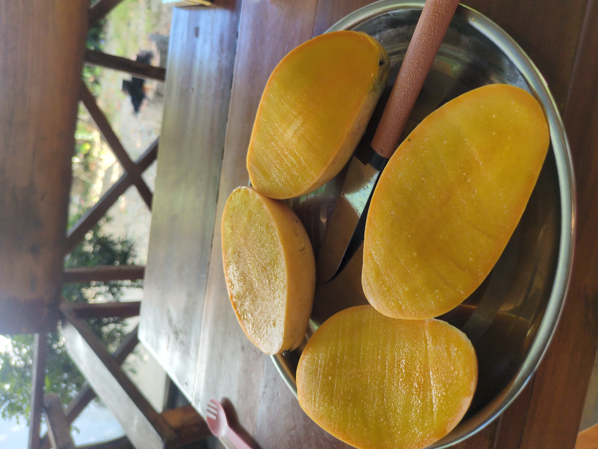 A high-angle shot shows a metal plate containing four mango halves and a knife, positioned on a wooden table. The fruit has a vibrant yellow hue and appears freshly cut. The knife has a brown handle and a metallic blade. In the background, a wooden structure with a view of greenery suggests an outdoor or semi-outdoor setting. A glimpse of what seems to be a plastic fork is visible near the edge of the table. Based on the style of the wooden structure and the tropical fruit, it is likely this image was taken in Phú Quốc, Vietnam.