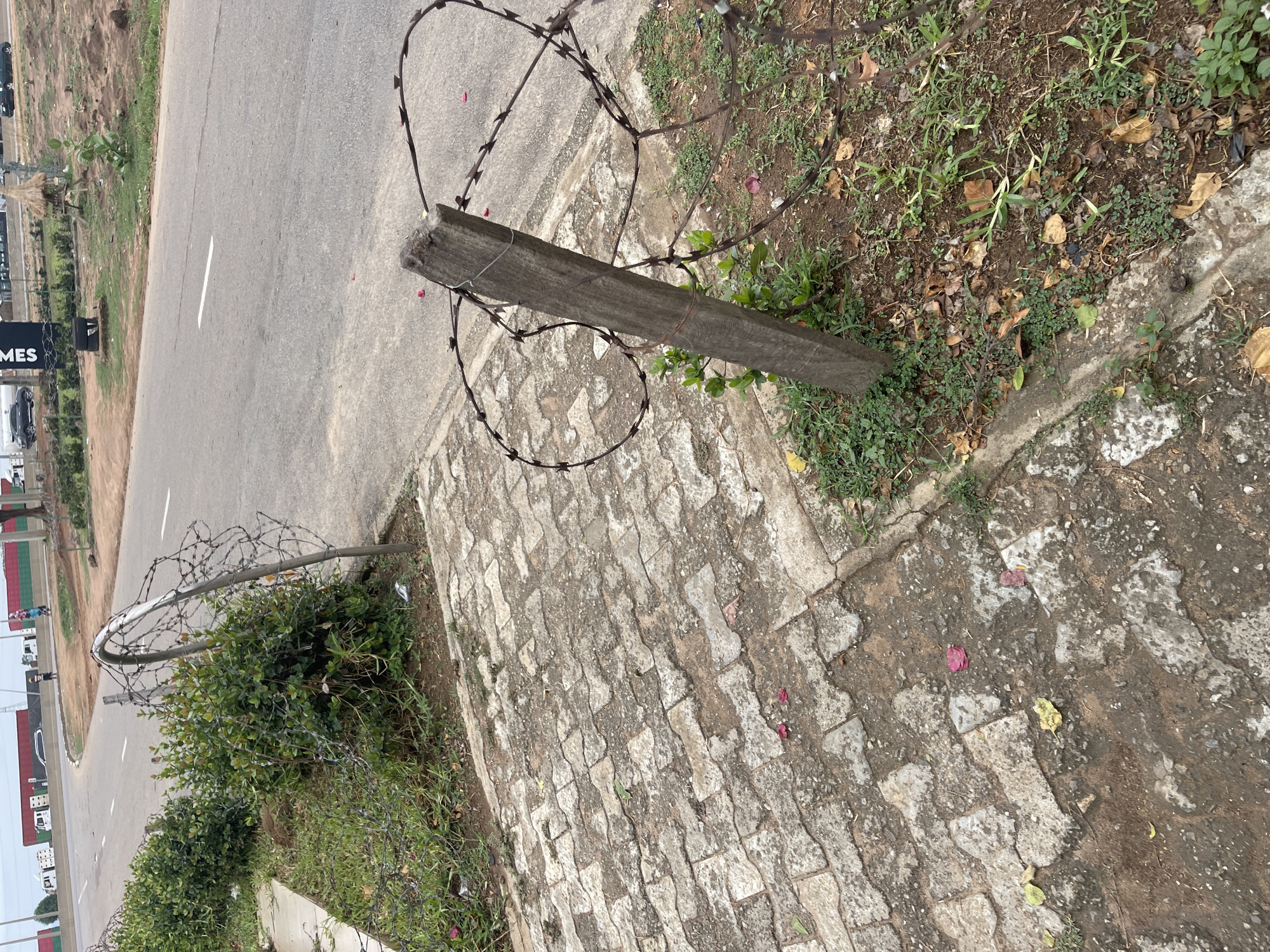 A photograph depicts a section of roadside in Gosa, Nigeria. A dilapidated wooden post is lying on the ground, entangled with rusty barbed wire. The immediate surroundings consist of paving stones, patches of grass, and soil. A road runs adjacent to the area. In the background, there are buildings and vehicles, suggesting an urban setting. No people are visible in the immediate foreground. The image is shot from a high angle.