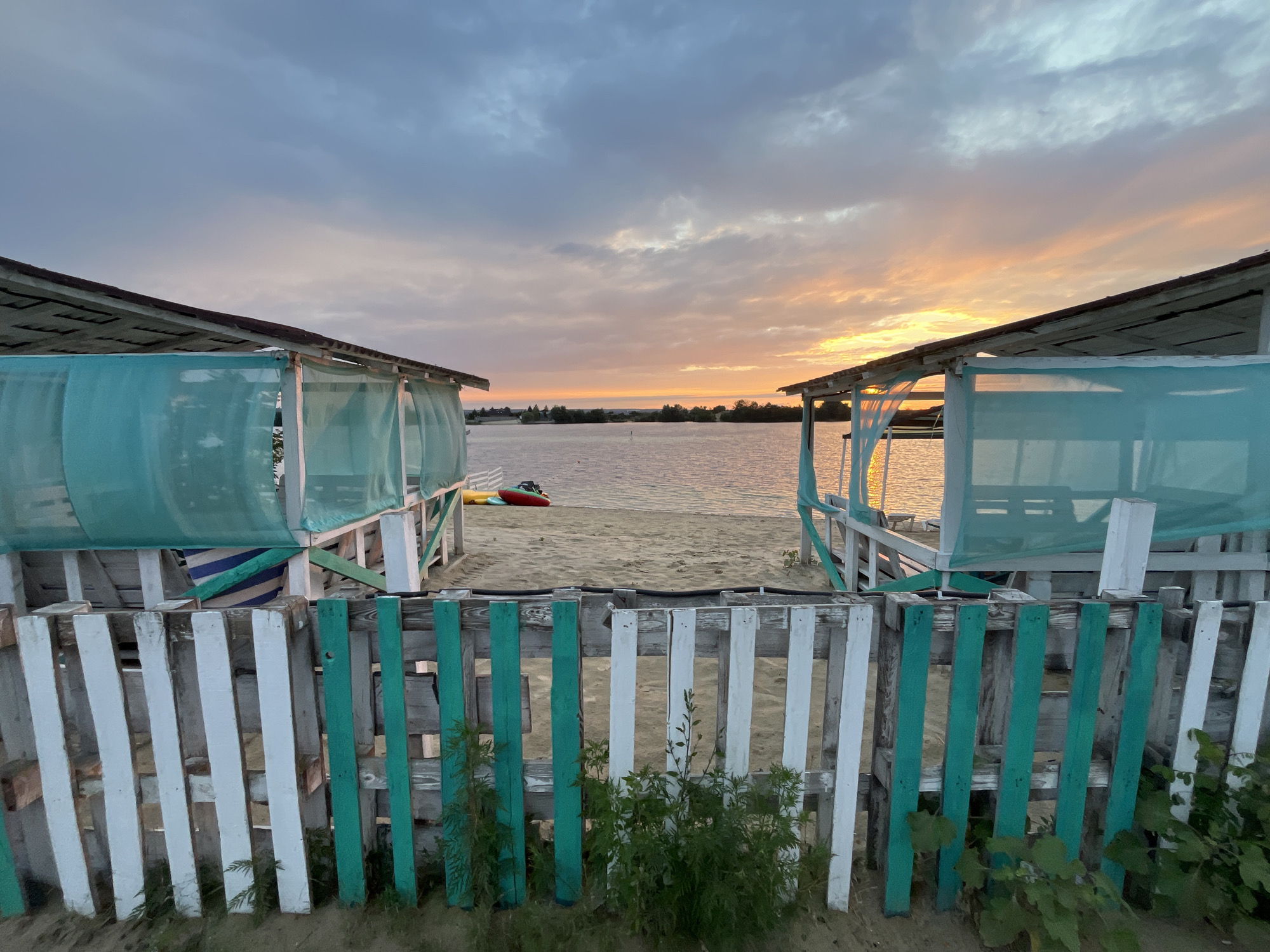 The image depicts a serene sunset scene at a beach on the Kharkiv Reservoir in Bezliudivka, Ukraine. Two white wooden structures with turquoise mesh curtains flank a sandy path leading to the water. A weathered fence, painted with alternating white and teal vertical planks, runs across the foreground, partially obscuring the view. On the sand, two kayaks are visible. The sky is painted with hues of orange, yellow, and grey, reflecting on the calm water. The environment suggests a tranquil evening at a recreational area.