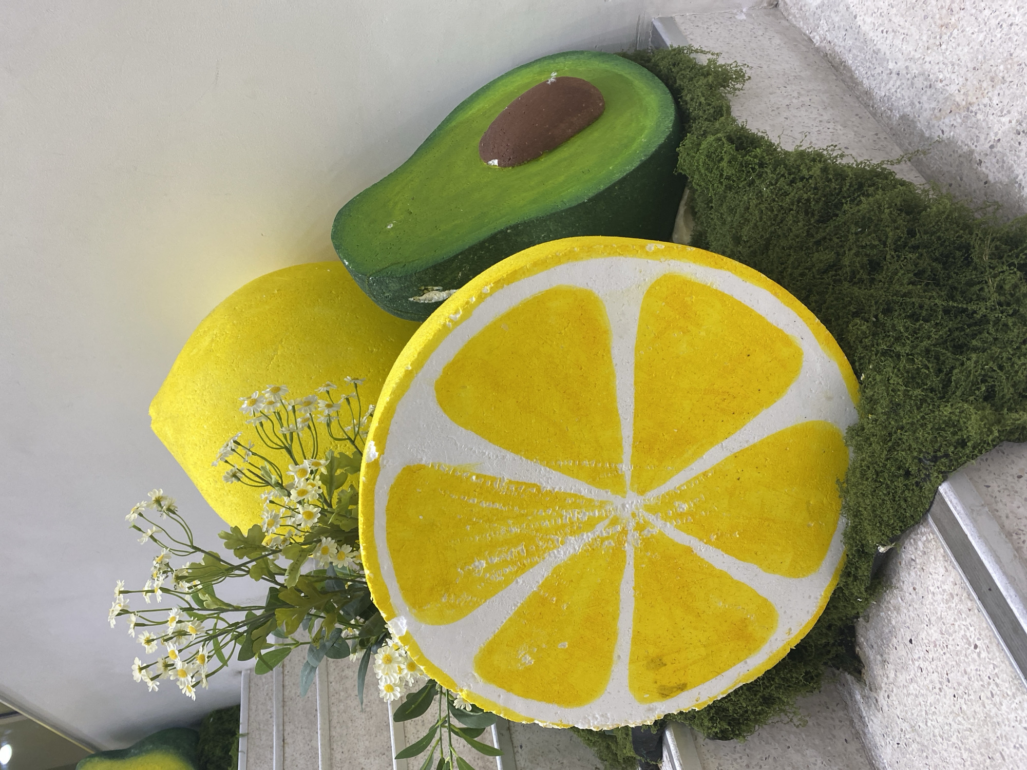 A decorative display of oversized fruit, including a whole lemon, a lemon slice, and a halved avocado, is placed on a light-colored, speckled staircase. Green moss covers the steps around the fruit. A bouquet of small white flowers with yellow centers and green leaves is situated behind the lemon slice and next to the whole lemon. The background wall is a plain, light color. This scene does not provide any specific indicators to determine its location within the city of Đà Nẵng, Vietnam.