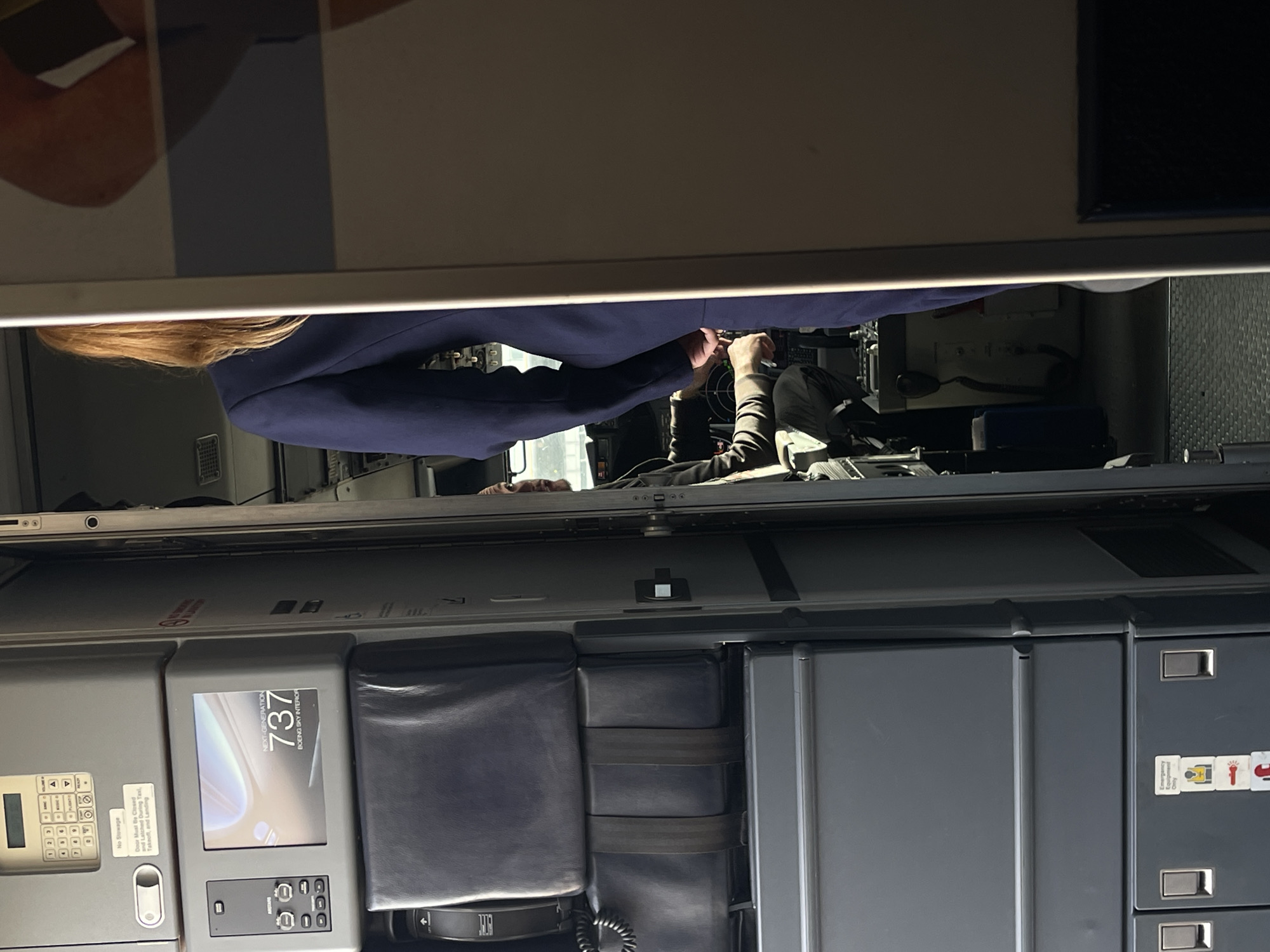 The rotated image, viewed upright, depicts the interior of an aircraft, specifically looking from the galley or forward cabin towards an open cockpit.
In the foreground, a person with long blonde hair, wearing a dark blue uniform jacket, stands at the threshold of the cockpit, observed from the back and side. Inside the cockpit, another individual, also in a uniform, is seated and appears to be interacting with the flight controls or other cockpit equipment. Faintly visible above the cabin features, a third person's face can be seen, possibly lying in a crew rest bunk.
The cabin area in the lower part of the frame features various control panels and storage units. On the left, there is a panel with a numerical keypad. Below it, a warning sign reads, "No Stowage" and "Door Must Be Closed and Latched During Taxi, Takeoff, and Landing." Adjacent to this, a screen prominently displays "NEXT-GENERATION 737 BOEING SKY INTERIOR" and "737," indicating the aircraft type. Below this screen is another control interface with buttons and a small display showing "00." To the right, several dark grey padded sections, possibly jump seats or equipment covers, are secured with straps. Further right, a series of grey cabinets or drawers are visible, one of which carries a label "Emergency Use Only" alongside icons for a flashlight and an oxygen mask. A coiled cable, likely for a crew interphone, is also present.
The scene suggests a period of pre-flight checks or preparations, with crew members active around the cockpit. The lighting is moderate, with some areas appearing dimmer than others. The overall setting is a modern aircraft interior. This media file was captured in Quart de Poblet, Spain.