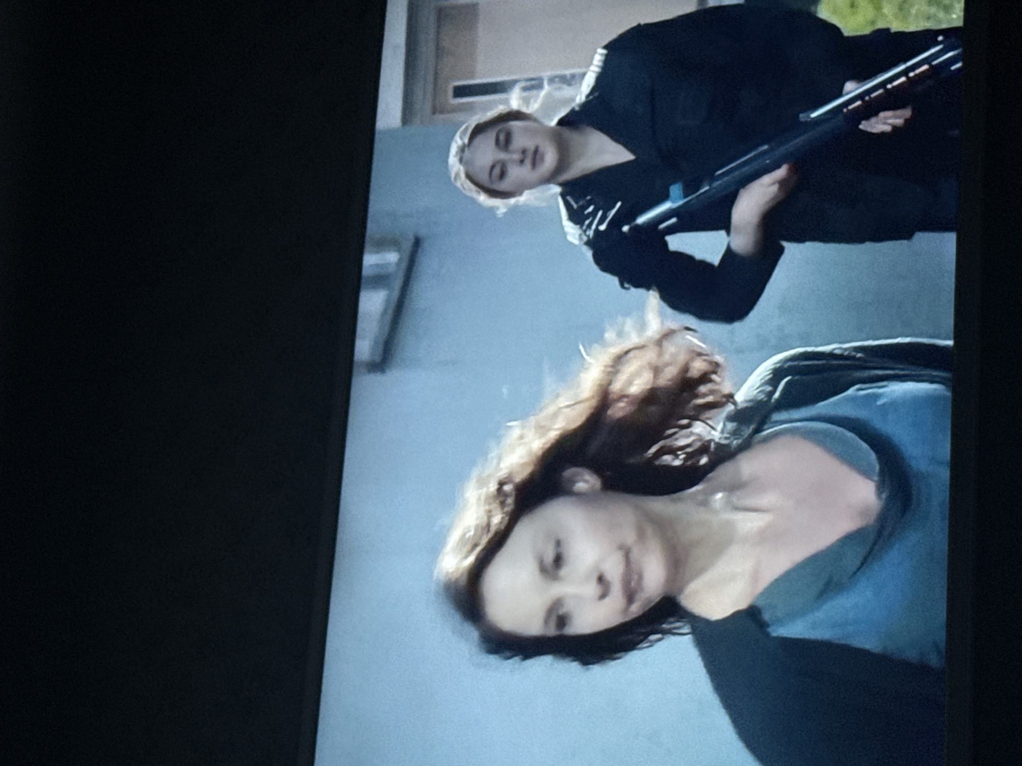 The image depicts two women outdoors. One woman is holding a long gun and is dressed in black. The other woman is in the foreground, wearing a blue top. The setting is adjacent to a light-colored building. Location is not discernible from the image.