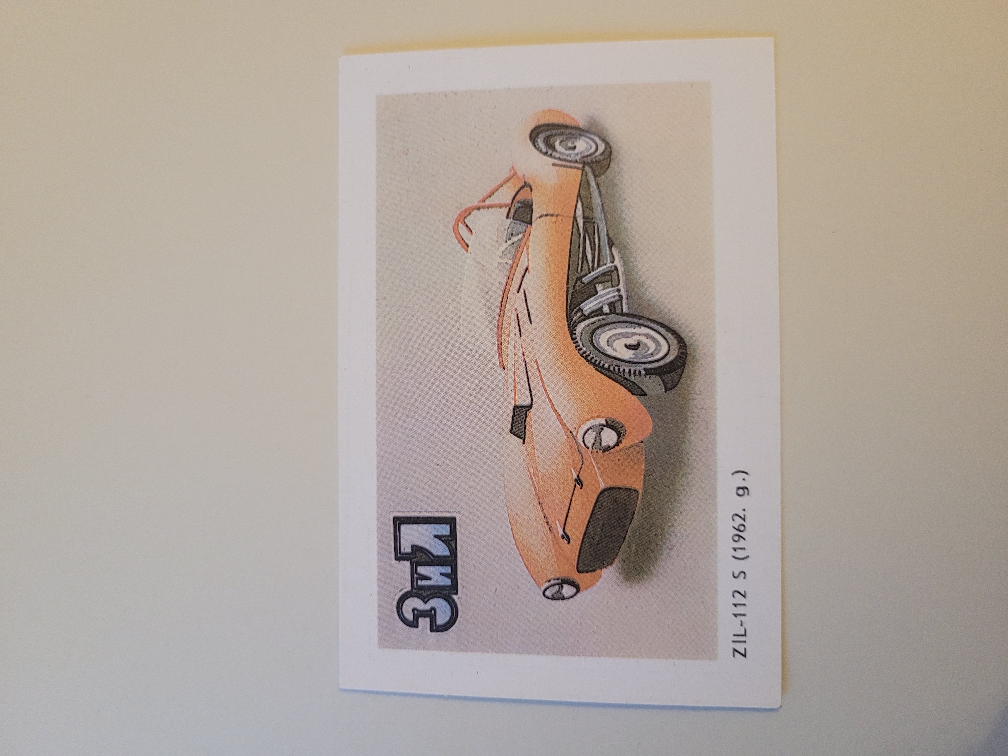 The image is a postcard featuring an illustration of an orange ZIL-112 S car from 1962. The car is depicted in a dynamic pose, with its front end angled towards the viewer. The illustration has a slightly faded, vintage aesthetic. In the upper left corner of the postcard, there is a stylized logo or inscription in a blue and white font that appears to be Cyrillic, possibly "ЗиЛ". Below the illustration, the text "ZIL-112 S (1962. g.)" is printed, identifying the car and its year of production. The postcard is set against a plain, neutral background. There is no indication of the city of Jūrmala, Latvia, in the image or its text.
