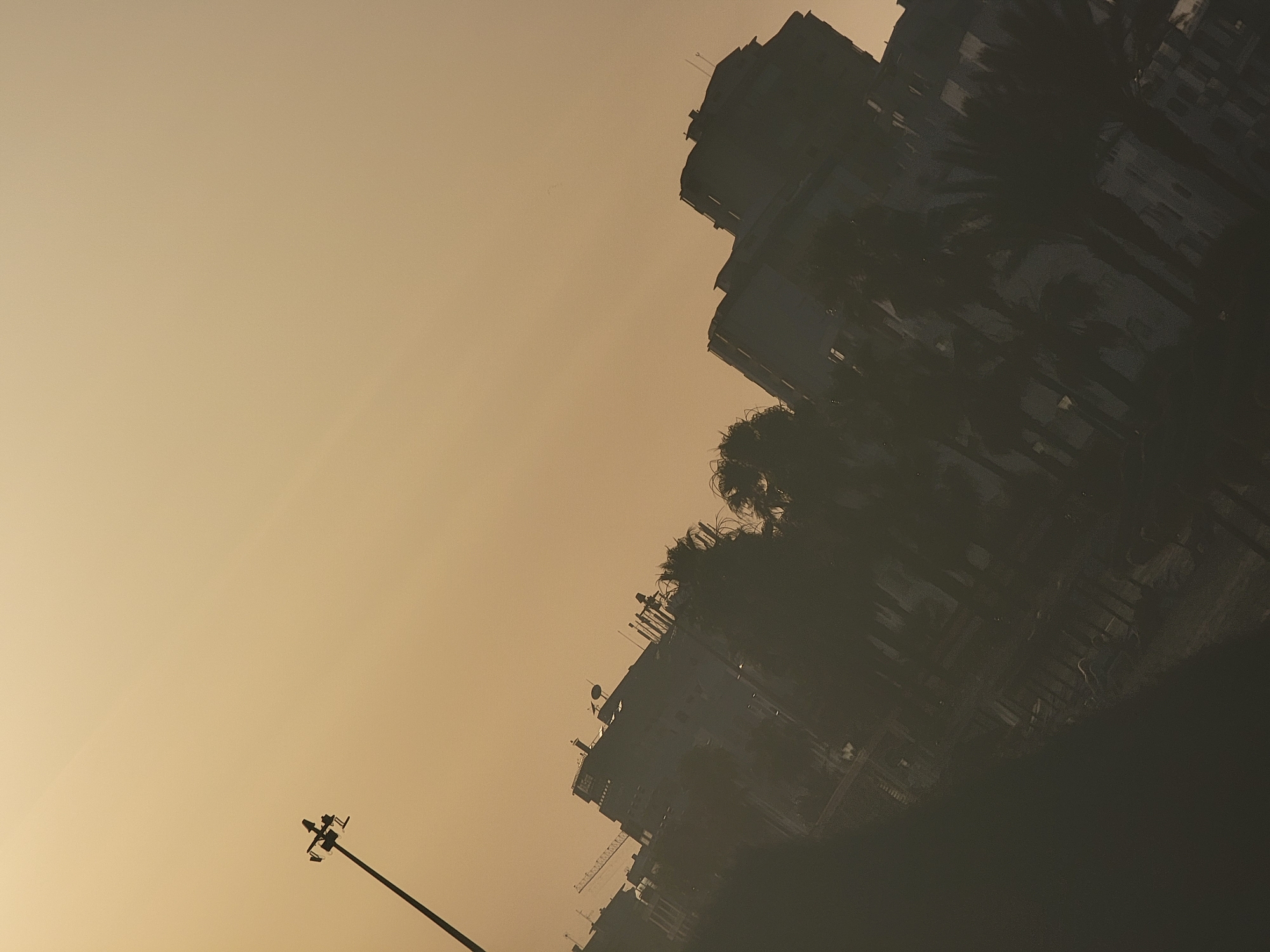 The image is a tilted, low-angle, silhouetted shot of a city skyline against a hazy, overcast sky. The overall color palette is muted, with tones of beige, brown, and dark grey, suggesting either a sunset or sunrise, or possibly smog.
In the upper left quadrant, a tall lamppost with multiple lights is visible, its silhouette stark against the sky. Extending diagonally across the frame from the lower left to the upper right are buildings and trees. The buildings appear to be multi-story structures, some with visible antenna or rooftop features. In the distance, a construction crane can be seen. Palm trees and other foliage are present in the foreground and middle ground, also silhouetted.
The scene appears to depict an urban environment, potentially Quarteira, Portugal, given the architecture and the presence of palm trees. The lighting suggests a low-light condition, obscuring fine details and creating a dramatic, somewhat mysterious atmosphere. No people or overt actions are visible. The image has a slightly grainy texture.