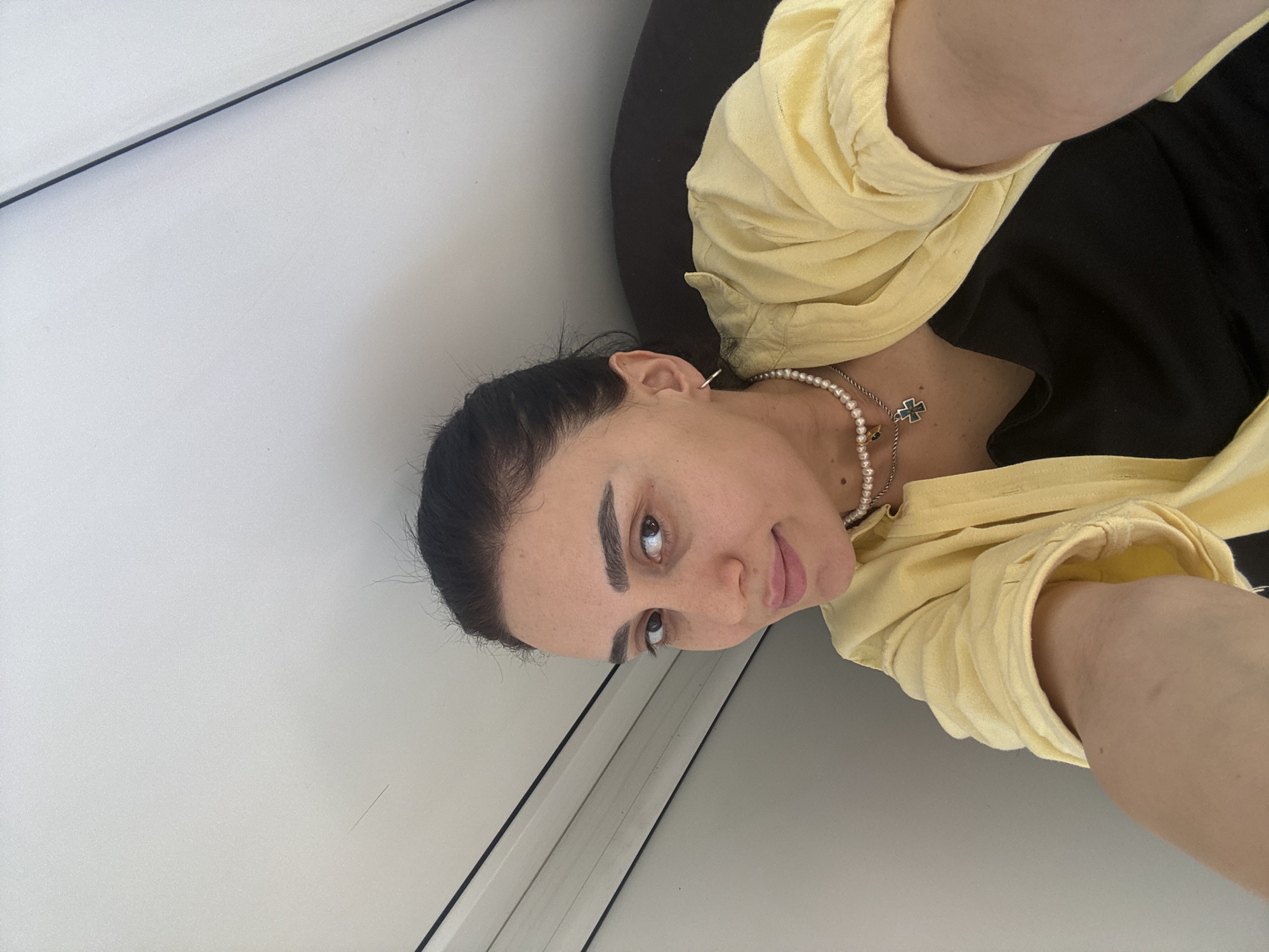 A woman with dark hair pulled back, wearing a yellow collared shirt over a black garment and a pearl necklace with a cross pendant, is looking up at the camera. She is indoors, against a backdrop of white walls and a window or door frame. The location is potentially Tbilisi, Georgia, but there is no definitive location identifier in the image.