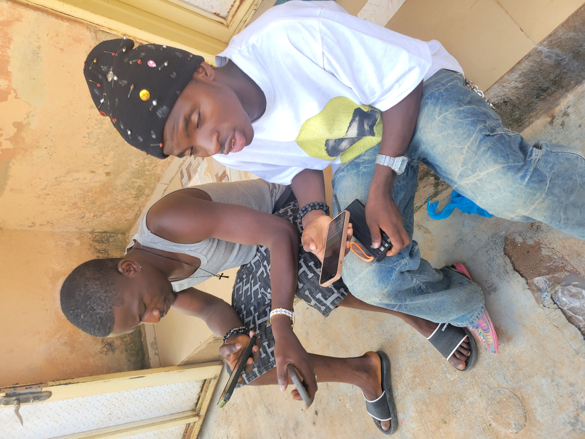 Two young men are sitting outdoors on a concrete step next to a beige wall. The man on the left is wearing a gray tank top, patterned shorts, and black flip-flops. He is holding two smartphones, one in each hand, and appears to be looking at the one in his left hand. He has a necklace with a cross pendant. The man on the right is wearing a black beanie adorned with colorful pins and beads, a white t-shirt with a yellow graphic, and distressed blue jeans. He is also holding a smartphone and speaking. He is wearing a large, silver watch on his left wrist and has a beaded bracelet on his right wrist. A blue plastic bag is visible on the ground between the two men. The environment suggests an urban setting, possibly an alleyway or the entrance to a building.