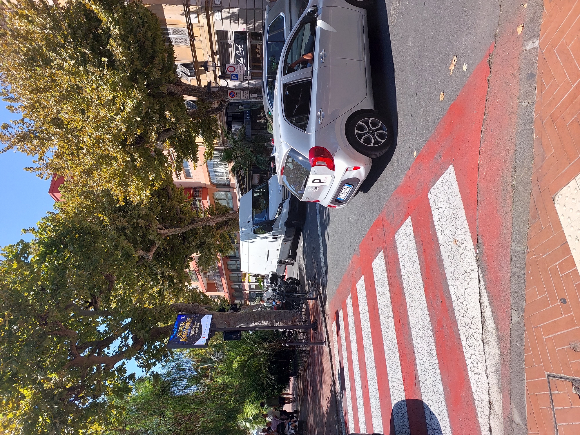 The image depicts a street scene in Ventimiglia, Italy, on a sunny day. In the foreground, a red and white striped crosswalk is visible, with a silver Kia Picanto car stopped at the intersection. The car has a "P" sticker on its rear window, indicating it is a learner driver. Behind the car, a white van is also stopped, and a motorcycle is parked nearby. In the background, several people are walking on the sidewalk, some with a stroller, and there are trees and buildings lining the street. A digital billboard displaying "SUPER KARAOKE" with "VENTIMIGLIA 3-4-5 SETTEMBRE" text is mounted on a pole. The architecture suggests a Mediterranean coastal town.