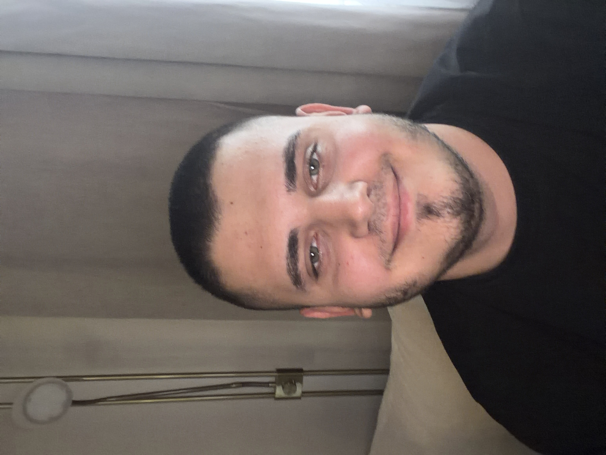 A young man with short dark hair and a beard smiles at the camera. He is wearing a black t-shirt. The background is a plain wall with a lamp and a curtain. The image was taken in a room in Sofia, Bulgaria.