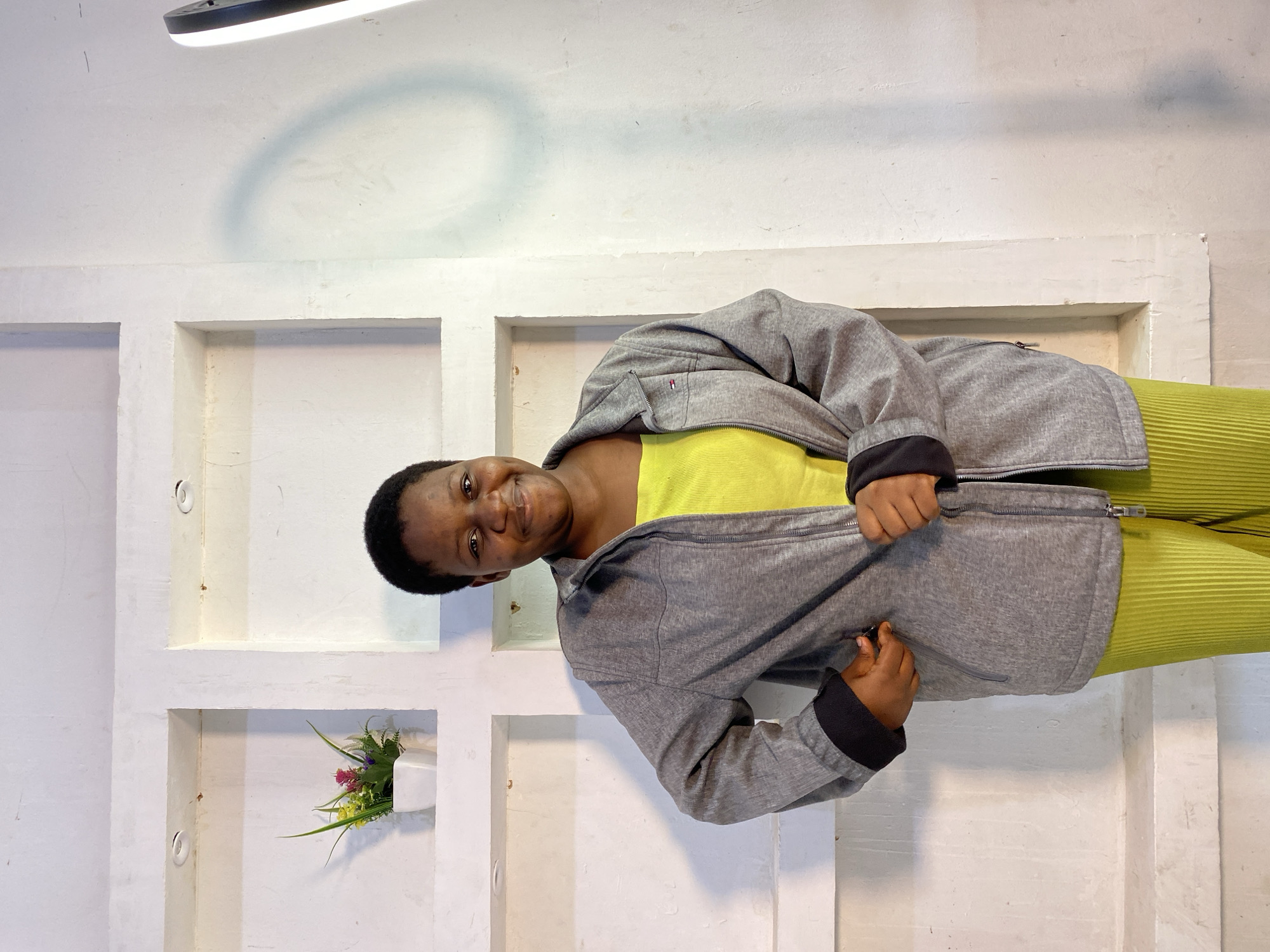 A young Black woman stands in front of a textured white wall with recessed square shelves. She has short, dark hair and is smiling at the camera. She is wearing a bright yellow top and a grey zip-up jacket. Her pants are a vibrant lime green and appear to be pleated. In the left-most shelf, a small white pot holds artificial pink and yellow flowers. The lighting casts a soft shadow of a circular object on the wall to her right. The scene is likely an indoor studio or a room with a decorative wall.