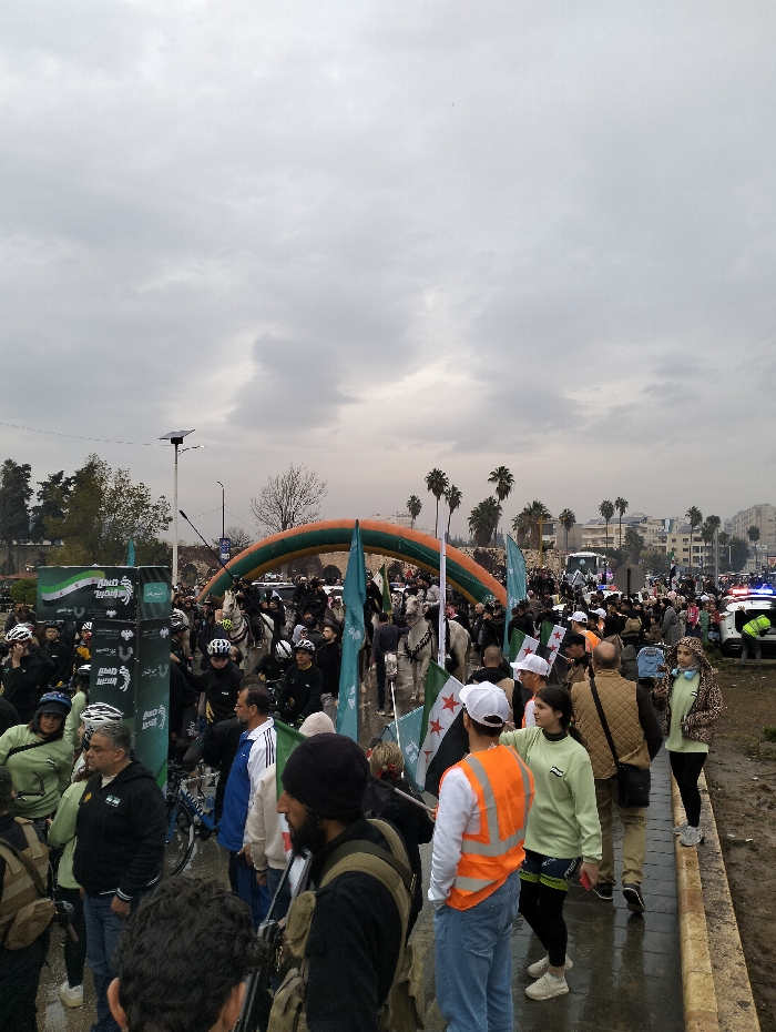 A crowd gathers under a cloudy sky, some holding flags of Syria. Police cars with flashing lights are visible in the background. An arch of green and orange balloons marks a starting or ending point. Individuals on horseback mingle within the group. Many people wear helmets. The ground appears wet and muddy. Trees and buildings are visible in the distance, along with palm trees.