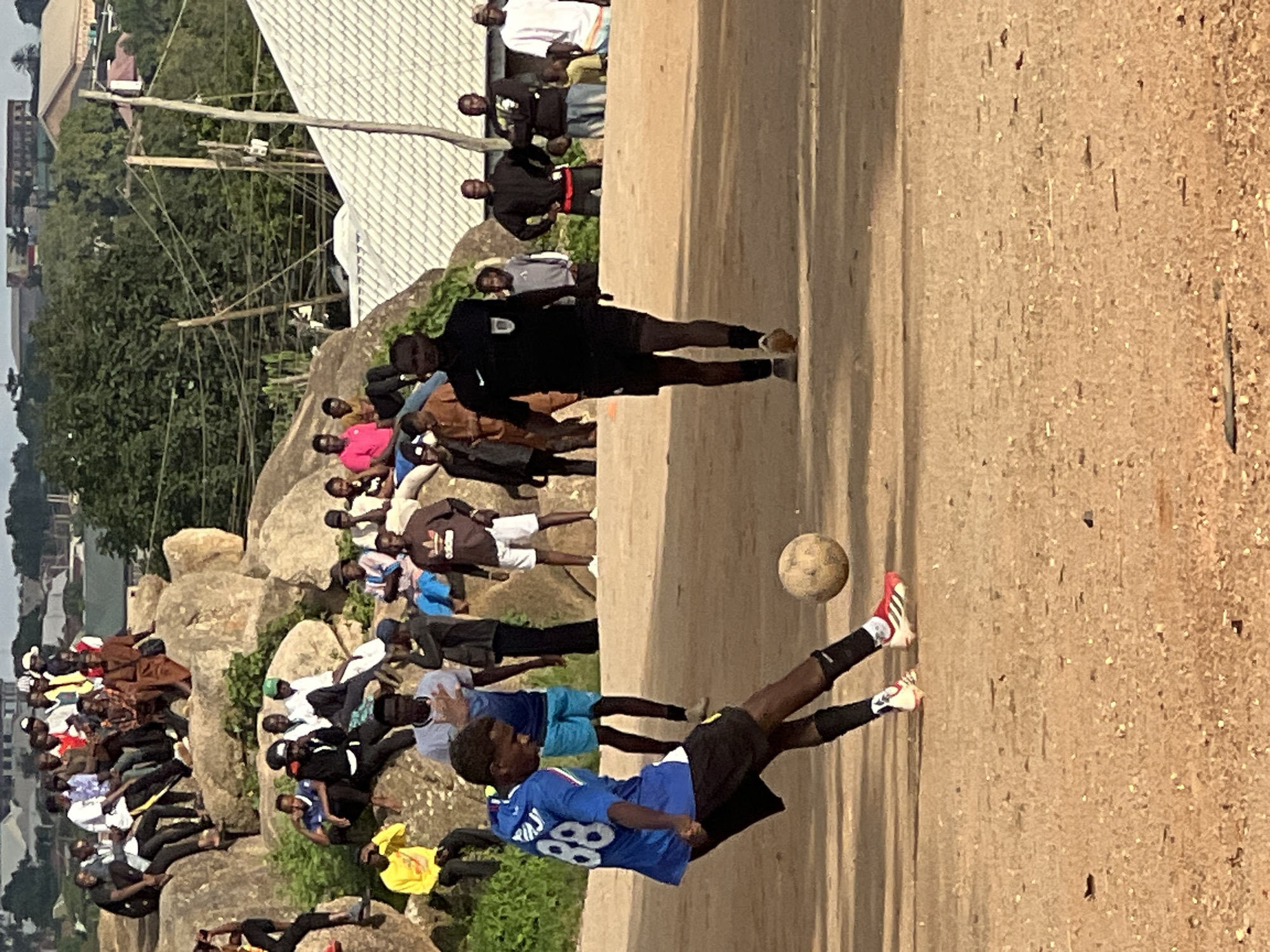 In Jos, Nigeria, a soccer game is underway on a dirt field. A young man in a blue jersey with the number 88 is kicking a worn soccer ball. He is wearing black shorts and black socks with red and white trim. The referee, dressed in a black shirt and black shorts, stands nearby. A crowd of spectators, many of them young men and boys, are gathered on and around large rock formations in the background. Some are seated on the rocks, while others stand. Buildings and trees are visible in the distance, suggesting an urban or semi-urban environment. The sky is overcast.
