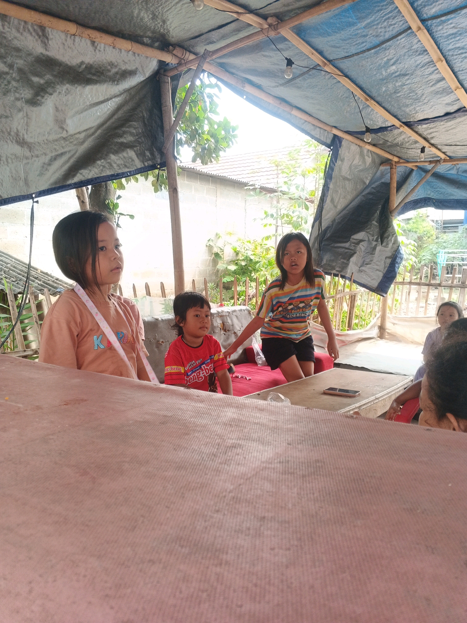 In the city of Kadu, Indonesia, a group of children are gathered under a makeshift shelter. The shelter is constructed with bamboo poles and draped with tarps, one a dark gray and the other blue, creating a dimly lit interior. The ground appears to be covered by a red mat or tarp.
In the foreground, a young girl with dark hair and wearing a light brown t-shirt with a pink and white strap across it is looking to the left. Beside her, a younger child in a red t-shirt is also looking forward. Further into the shelter, a girl with dark hair is wearing a striped t-shirt and black shorts, standing and looking towards the camera. Another child, wearing a light purple shirt, is seated on the right side of the frame, partially obscured. A table is visible in the center of the shelter, with a mobile phone resting on it. The background reveals a glimpse of buildings and vegetation, suggesting an outdoor, possibly rural or semi-urban, setting. The overall atmosphere appears casual, with the children seemingly engaged in some activity or simply present within the sheltered space.