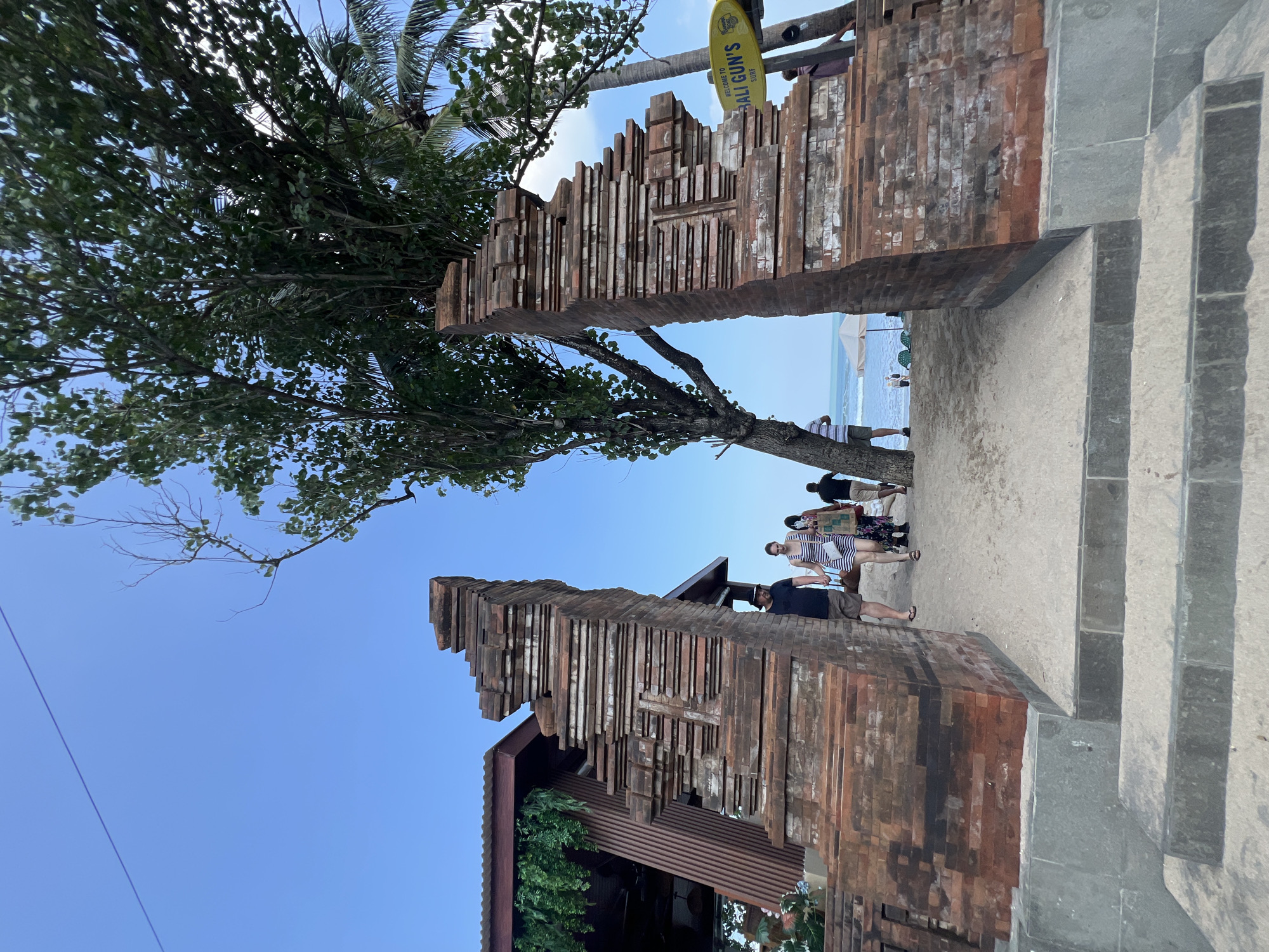 In Kuta, Indonesia, a tall, weathered brick gateway stands open on a sandy path leading to a beach. The gateway's ornate, layered structure is partially obscured by the dense foliage of a large tree, with palm fronds visible in the background. A couple, holding hands, walks through the gateway toward the beach. The man wears a black t-shirt and shorts, while the woman is in a striped dress. Behind them, other individuals walk in the same direction. To the right of the gateway, a yellow surfboard sign reads "WELCOME TO BALI GUN'S SURF." The ocean is visible beyond the sand, with a few beach chairs and umbrellas set up. The sky is clear and blue, with a thin, purple line possibly indicating a wire or string stretched across the frame. To the left, a building with a sloped, reddish-brown roof and vertical wooden siding is partially visible, adorned with hanging green foliage.