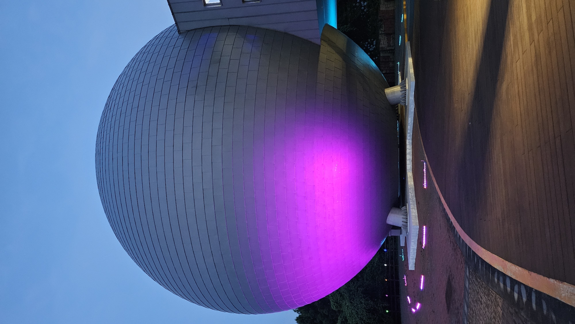 The image depicts the exterior of the Jeju National University Museum of Art, a spherical building illuminated with vibrant pink and blue lights. The building's metallic facade is composed of horizontal panels, and it rests on three large, claw-like supports. A walkway paved with wooden planks and brick patterns curves around the building, lined with small, glowing pink lights. Trees are visible in the background, and the sky is a clear, deep blue, suggesting it is dusk or early evening.