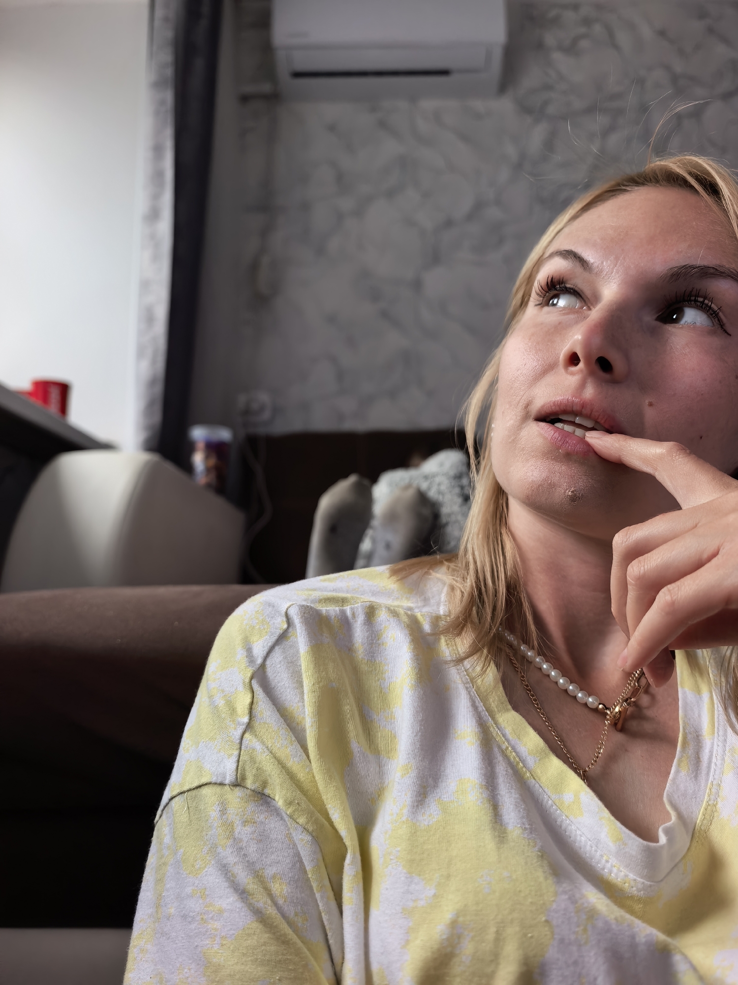 A close-up, low-angle shot features a young blonde woman, likely in her 20s or 30s, captured indoors in Sterlitamak, Russia. She is looking upwards and slightly to her right, with a pensive or thoughtful expression. Her right index finger gently touches her lower lip, a common gesture of contemplation. Her hair is light blonde, and her eyes are directed off-camera. She is wearing a casual yellow and white tie-dye patterned t-shirt, and accessorized with a pearl necklace and a delicate gold chain necklace with a small charm.
The background shows elements of a modern home interior. An air conditioning unit is mounted on a wall with a distinctive grey and white marble-like pattern. Dark curtains are visible on the left. Furniture includes a brown sofa or armchair with a lighter-colored armrest. On a surface behind the sofa, a red cup and a transparent container filled with snacks are visible. Further in the background, obscured by blur, are other pieces of furniture and a light-colored object. The lighting in the room suggests it is daytime, creating a soft, ambient glow. There is no visible text in the image.