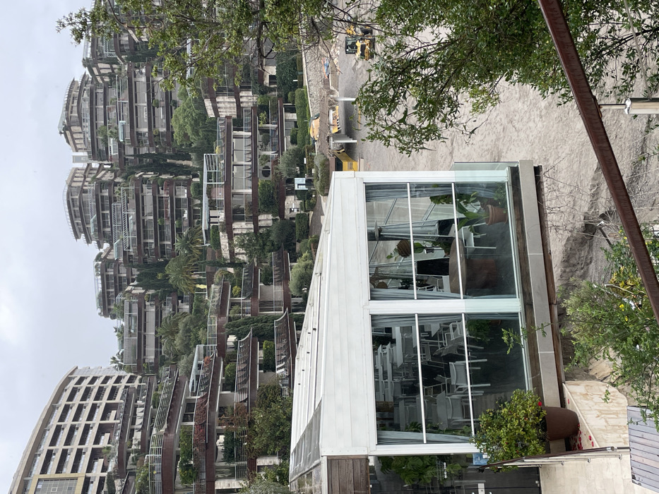 The image displays an elevated view of the Dukley Gardens residential and resort complex in Budva, Montenegro, under an overcast sky.
In the immediate foreground, a dark brown metal railing with silver uprights partially obstructs a view of green foliage. To the left, a brown pot containing a green plant rests on a light-colored paved surface, adorned with scattered red petals. Dominating the central foreground is a white-framed, glass-enclosed structure, identified as a dining area due to visible arrangements of multiple white square tables and matching chairs within. This structure features a translucent, multi-panel roof and a glass railing along its perimeter. Inside, two large potted plants and a cylindrical brown heater are visible.
The midground presents a broad, light-colored expanse of sand or gravel, indicative of a beach or an active construction/landscaping zone. A bright yellow compact excavator is positioned on this surface toward the right, oriented to the right. To the left of the excavator, a partially obscured structure with a thatched roof and yellow materials suggests a beach bar or shaded area under construction or maintenance.
The background is characterized by the distinctive architecture of the Dukley Gardens complex, comprising numerous multi-story, terraced residential buildings ascending a green hillside. These modern structures feature dark-toned, curvilinear balconies and facades, extensively integrated with green landscaping, including various trees, shrubs, and palm trees. A large, curving, lighter-colored building is prominent on the far left of the complex. The sky above is uniformly grey and cloudy. No individuals are visible, and no active movements are depicted in the scene.