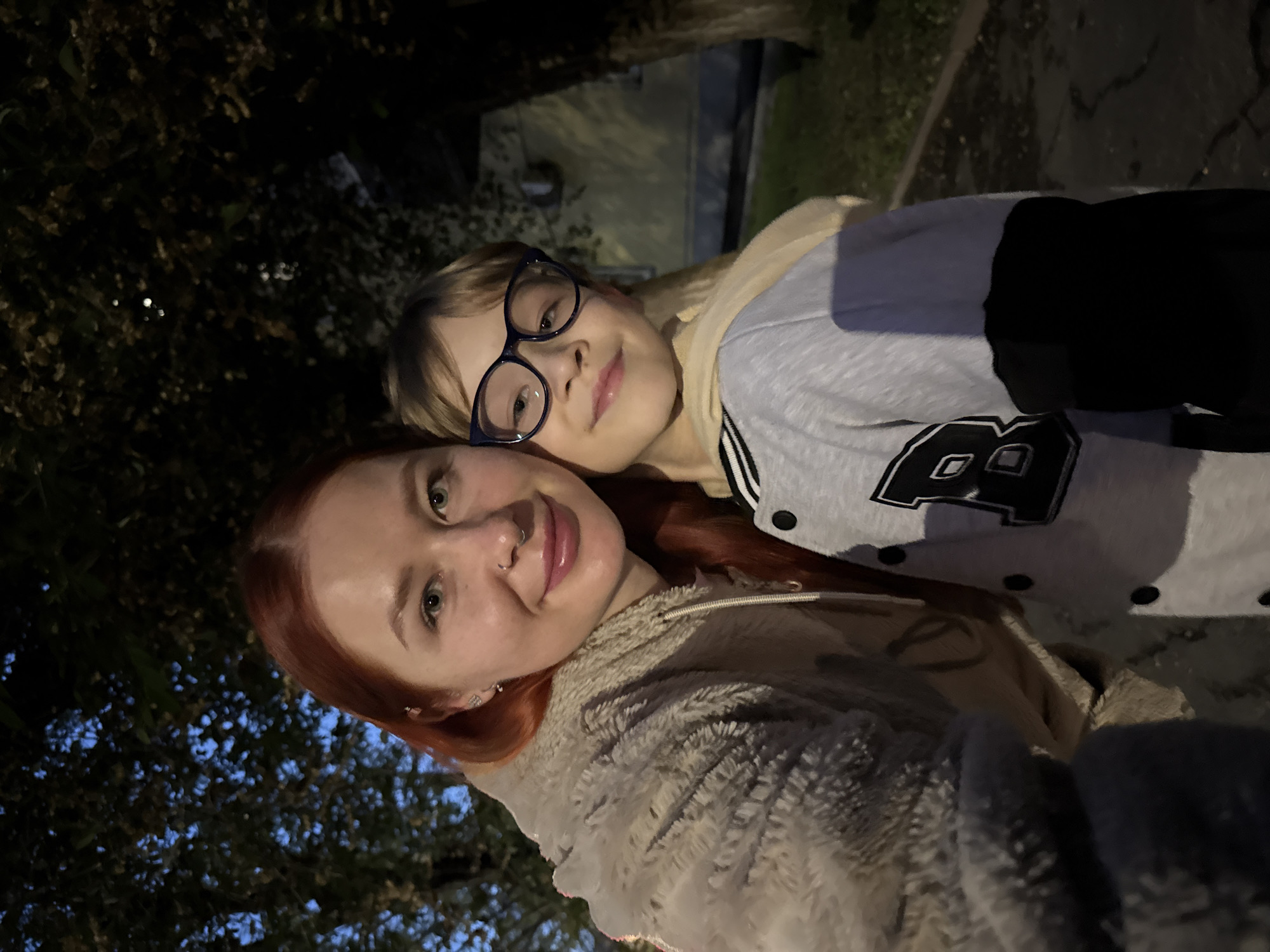 A selfie of two people at night. A woman with red hair and a nose piercing is wearing a fuzzy jacket and holding the camera. Standing behind her is a child with blonde hair and glasses, wearing a gray and black jacket with a large letter "A" on it. The background is dark, but trees and a building can be seen. The location is likely outdoors, possibly in a park or residential area. There are no indicators of the location being Rostov-on-Don.