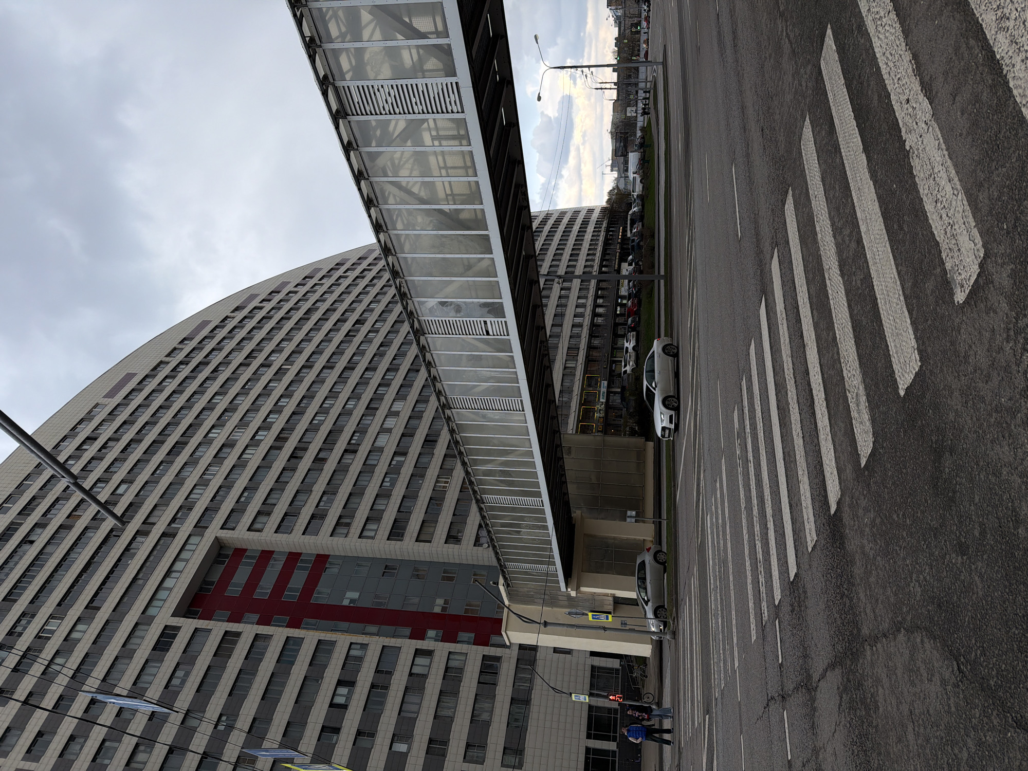 The image shows an urban scene in Khimki, Russia, featuring a large, curved building connected to another building across a street by an enclosed pedestrian bridge. The building is predominantly light gray with rows of windows and a red-accented section. The bridge is constructed of metal and translucent panels. Below the bridge, at street level, a crosswalk is visible on a gray asphalt road with white lane markings. A few cars are parked near the buildings. Two people appear to be standing near a traffic light at the crosswalk. The sky is overcast.