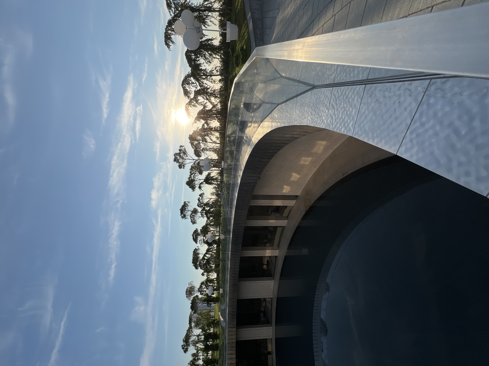 The image depicts a view overlooking a circular, dark body of water. The water is surrounded by a curved, modern architectural structure with concrete pillars and glass-enclosed rooms. A sleek, metallic railing with a glass barrier runs along the edge of the elevated walkway from which the photo is taken. The railing is illuminated by the warm glow of the setting sun, which is visible in the background, peeking through a line of tall, slender trees. The sky is a clear blue with wispy white clouds. In the distance, more trees and some white, abstract sculptures resembling large balloons in pots can be seen. The overall setting suggests an outdoor public space or park. This scene is located in Krasnodar, Russia, at the "Innopolis Park" or "The Flowers" park, a contemporary architectural complex designed by architect Sergey Skuratov.