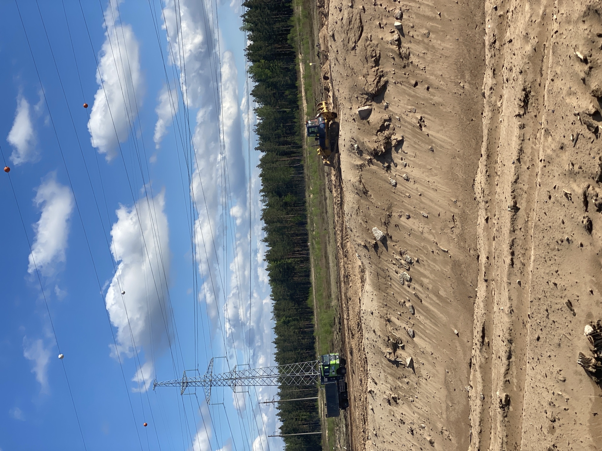 The image shows a construction site in Luzhitsy, Russia. A dirt pit or excavation area is visible on the right side of the frame. A bulldozer is positioned along the edge of a treeline. Further down, a green truck with a trailer is situated beside an electricity pylon supporting high-voltage power lines. Orange marker balls are attached to the power lines. The sky is blue with scattered white clouds.