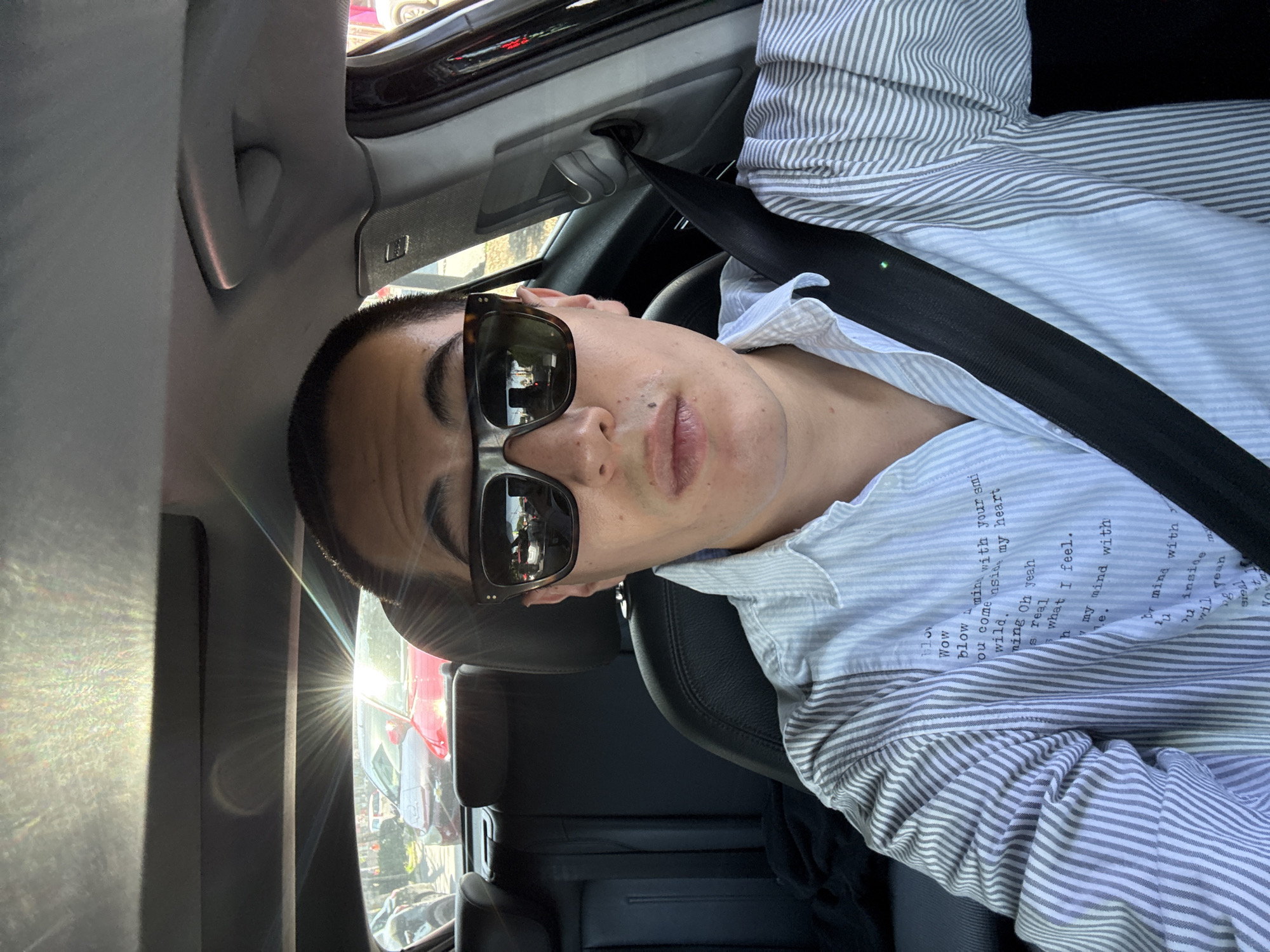 A young man is captured in a selfie-style photograph, seated in the passenger seat of a car, likely in Puebla, Mexico, on a bright, sunny day. He is seen from the chest up, looking directly towards the camera with a neutral expression.
He wears a stylish light blue and white striped button-down shirt. The light blue sections of the shirt are adorned with printed text in a handwritten font. Visible phrases from the text include "Wow," "blow," "my mind with your smile," "my heart," "You come inside," "wild," "Oh yeah," "is real," and "what I feel."
His face is partially obscured by a pair of dark sunglasses, which have brown tortoiseshell frames with metallic silver accents on the sides. Reflections in the sunglass lenses show a vibrant outdoor street scene with other vehicles and structures, indicative of an urban environment. A strong lens flare from intense sunlight is prominent on the upper left side of the image, suggesting the sun is shining directly into the car.
A black seatbelt is fastened across his chest and left shoulder. The car's interior features dark-colored leather seats, a dark dashboard area, and light grey ceiling trim. Details such as a grab handle and a small speaker grille are visible on the ceiling panel. The overall scene conveys a casual moment during a daytime journey.