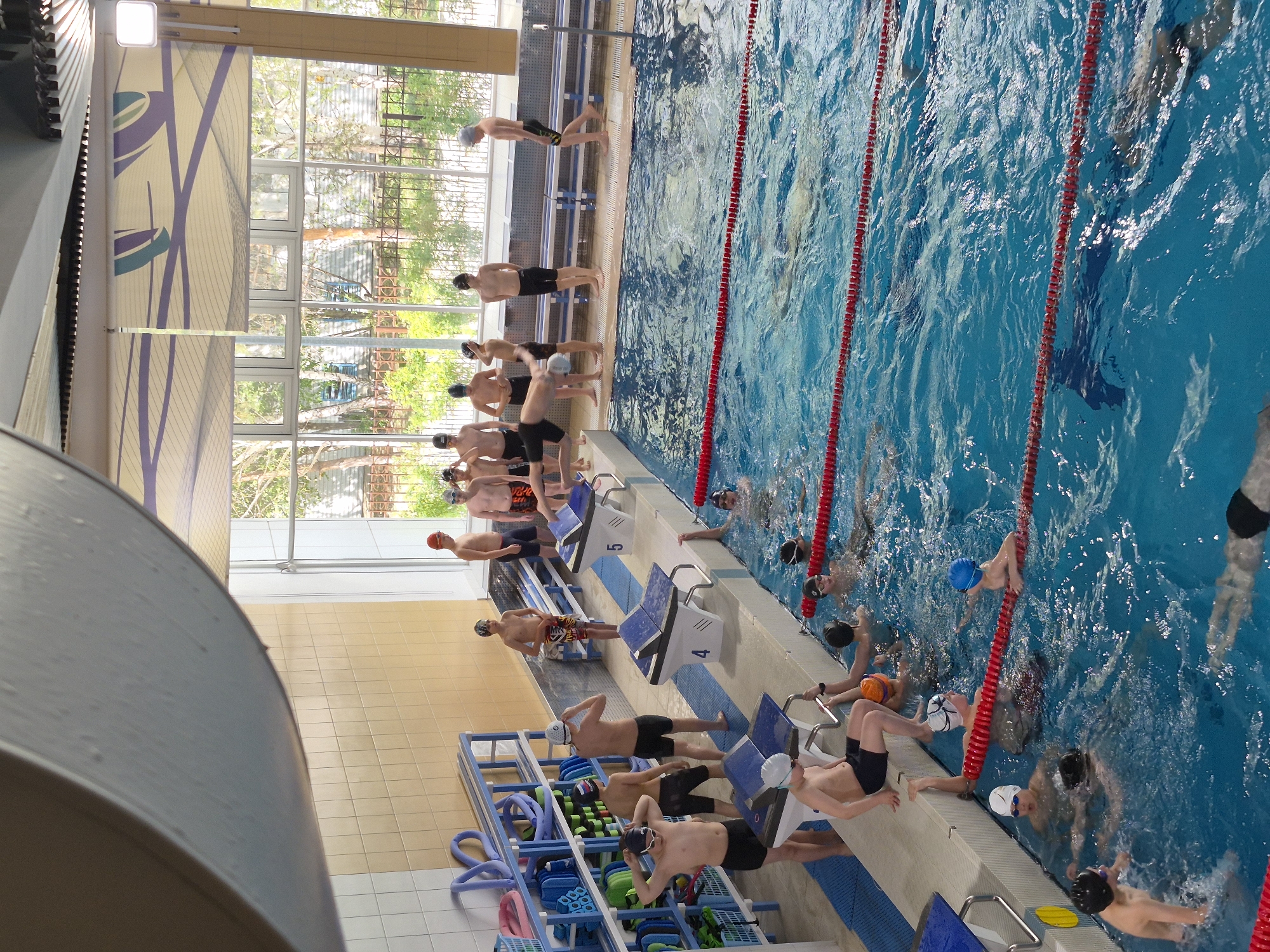 The image depicts an indoor swimming pool, possibly in Tobolsk, Russia given the information provided. A group of young male swimmers are present, some standing on starting blocks numbered 4 and 5, while others are already in the water. The pool is divided by red lane markers. Some swimmers in the pool are holding onto the lane markers. Several individuals are wearing swim caps and goggles. Adjacent to the pool is a yellow and blue tile-lined area with blue storage racks holding pool equipment like kickboards and pull buoys. Large windows allow natural light into the pool area. The building’s architecture features a modern design with a white ceiling and geometric patterns.