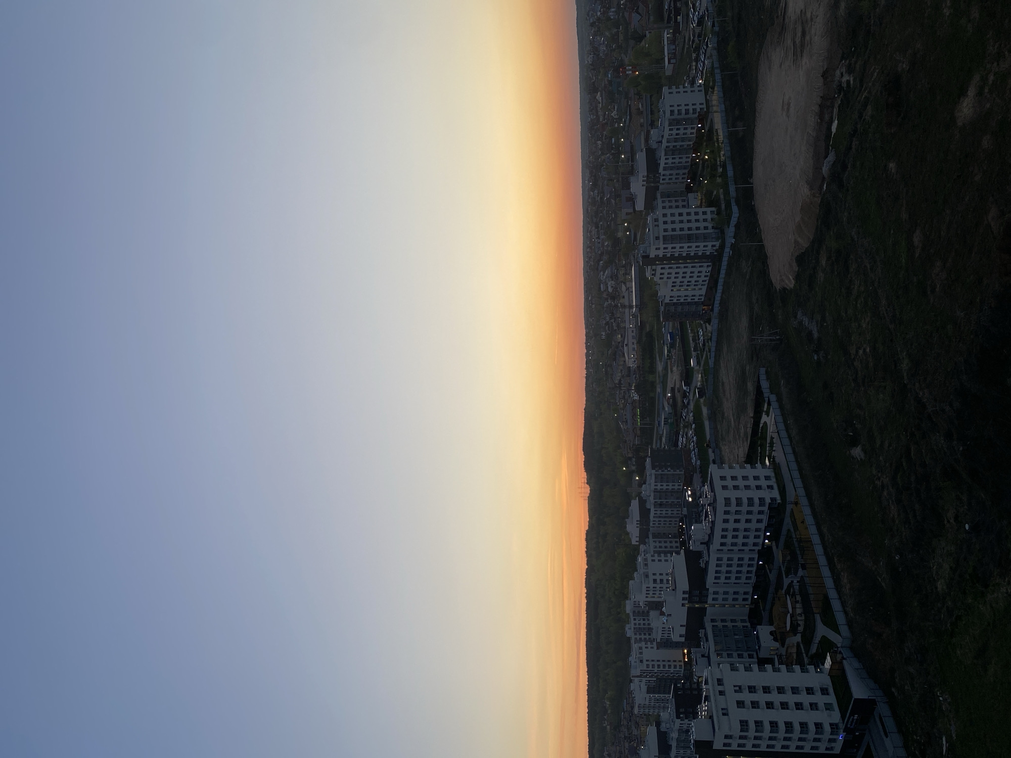 The image depicts a cityscape at sunset, likely in Kazan, Russia, judging by the urban layout and architectural style. The sky transitions from a light blue on the left to a warm orange-yellow gradient near the horizon. The cityscape includes multi-story apartment buildings with visible windows, interspersed with trees and green spaces. There's a construction site or a large patch of bare earth visible on the right side of the frame, alongside a darker, grassy area. Streetlights illuminate portions of the area near the buildings, suggesting dusk or early evening.