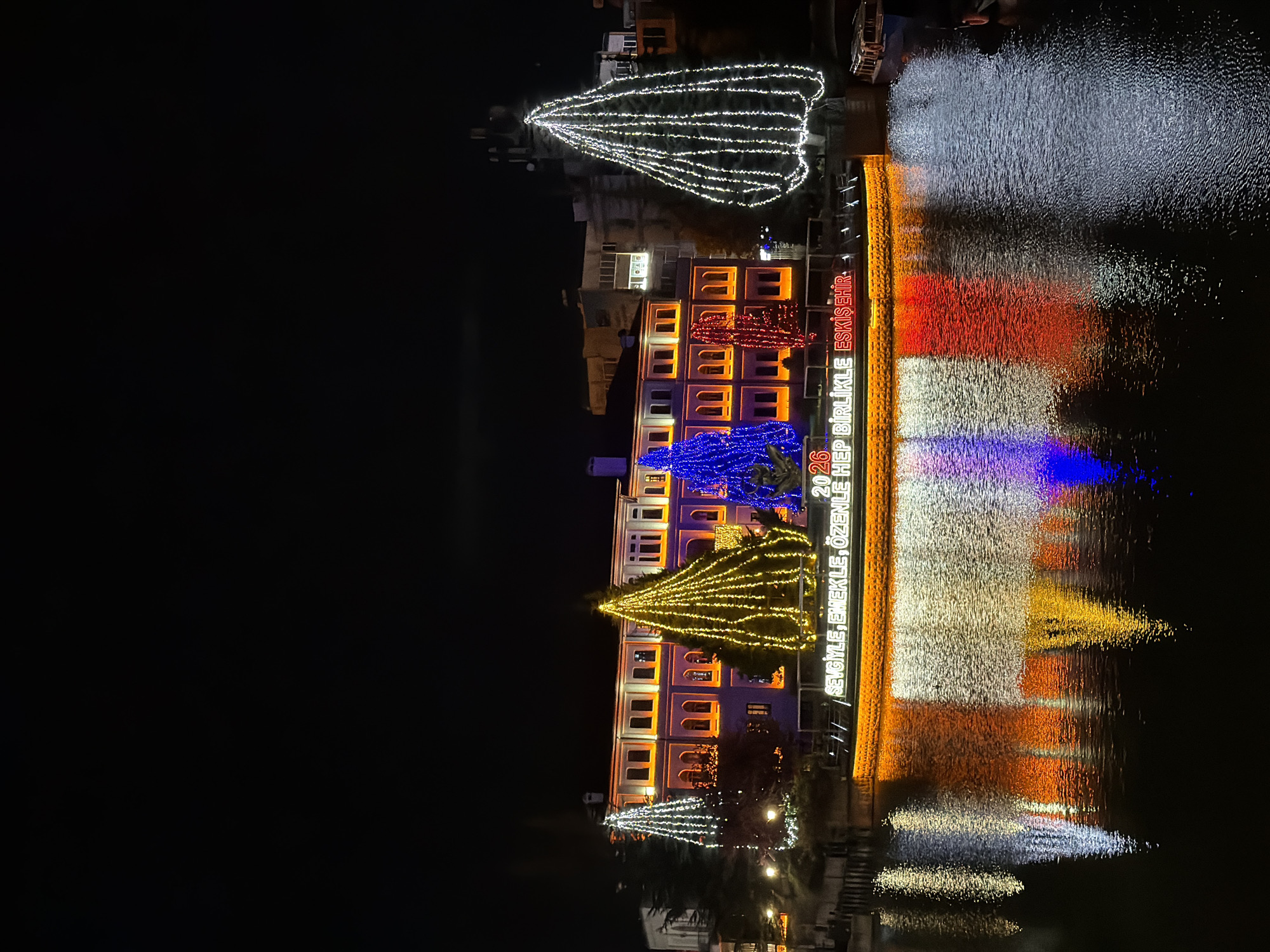 The image captures a vibrant night scene in Eskisehir, Turkey, showcasing a building and its surroundings adorned with an elaborate light display. The main subject is a multi-story building, possibly a government or municipal building, illuminated with colorful lights. Its windows and architectural details are outlined in warm orange and white lights, with a section featuring red lights.
In front of and around the building, several large, conical tree-like structures made of string lights are prominent. These installations vary in color, with one large one appearing in bright white lights, another in deep blue, and a third in golden yellow, suggesting festive decorations. Another smaller white light structure is visible further down the bank.
The lights from the building and the illuminated structures are vividly reflected in the calm body of water in the foreground, creating striking horizontal bands of white, red, blue, and yellow across the surface. This reflection enhances the overall magical and festive atmosphere of the scene. The time of day is clearly night, with the dark sky providing a stark contrast to the brilliant lights.
Visible text on the building, illuminated in white, reads "2026", "HEP BİRLİKLE ESKİŞEHİR", and "SEVGİYLE, EMEKLE, ÖZENLE H...". These phrases, particularly "ESKİŞEHİR", confirm the location context. The display suggests a public celebration, possibly for a new year or a significant city event, emphasizing community and dedication as indicated by the text.