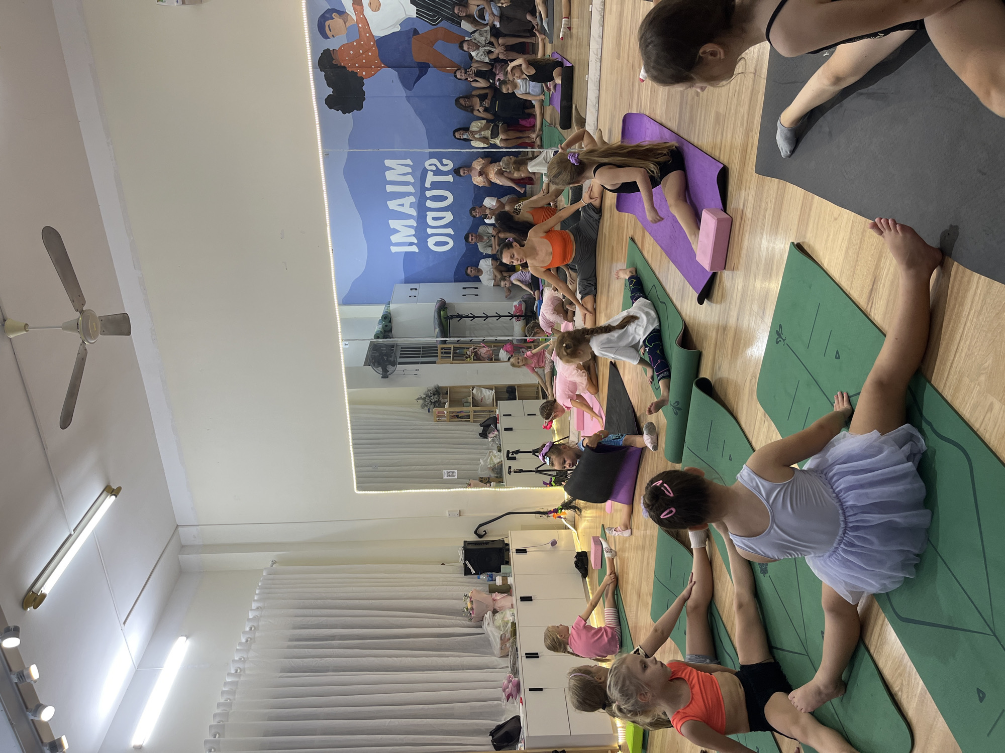 In a brightly lit studio with wooden floors and white walls, a group of young girls in athletic wear are participating in a yoga or dance class. They are positioned on green and purple yoga mats, many in a seated split position. A female instructor, wearing an orange top and grey shorts, is in the foreground, also demonstrating a split. In the background, a mirror reflects the studio, showing more participants and spectators seated along the wall. A mural with a blue mountain landscape and text that reads "MAIM STUDIO" is visible on the back wall. The scene suggests a children's activity session in a professional studio setting. The location is identified as Bac Nha Trang Ward, Vietnam.