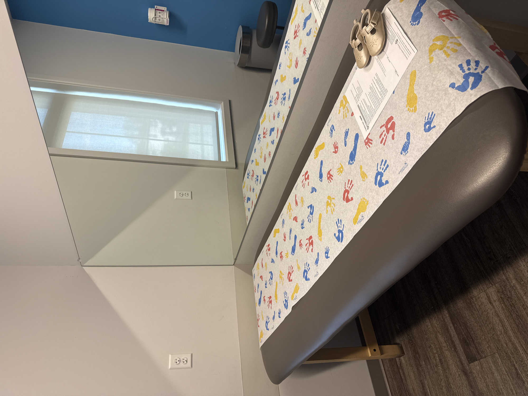 The image shows a medical examination room, likely in Hilliard, Ohio, based on the location constraints. The room has light gray walls, a blue ceiling, and dark wood-like flooring. A rectangular window with white blinds is visible on the left wall. The examination table is the central object. It is covered with a white paper sheet printed with colorful hand and foot prints. Two small, gold-colored baby shoes and a medical document lie on the exam table. An examination light is mounted on the wall near the top of the table. Two electrical outlets are visible on the left wall, and another closer to the floor. The image appears to be taken in a medical office or clinic specializing in pediatric care. There are no people present in the image.