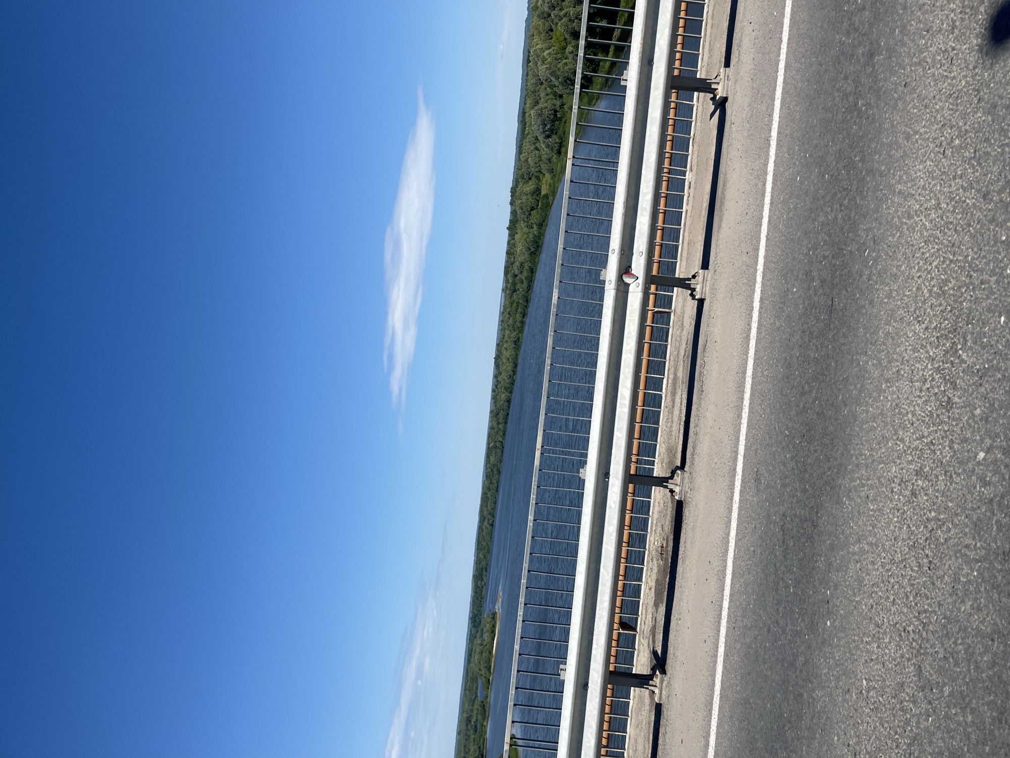 The image shows a view from a bridge over a wide river. The sky is clear and blue with a few white clouds. The river is a deep blue color and is bordered by lush green trees on both sides. A metal guardrail runs along the side of the bridge, with an asphalt road in the foreground. The location is identified as the city of Yadrin, Russia. There are no people or animals visible in the image. The scene is tranquil and showcases a natural landscape.