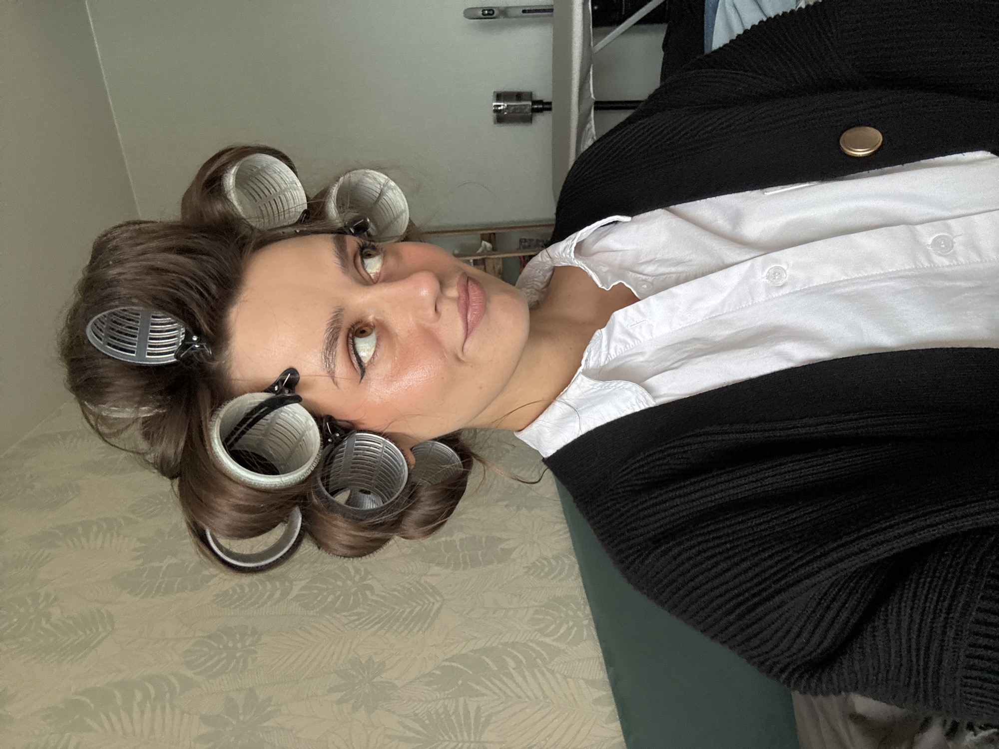 A woman with brown hair in curlers is lying on a surface with a leafy pattern. She is wearing a white collared shirt with a buttoned black cardigan over it. The location cannot be determined based on the image.