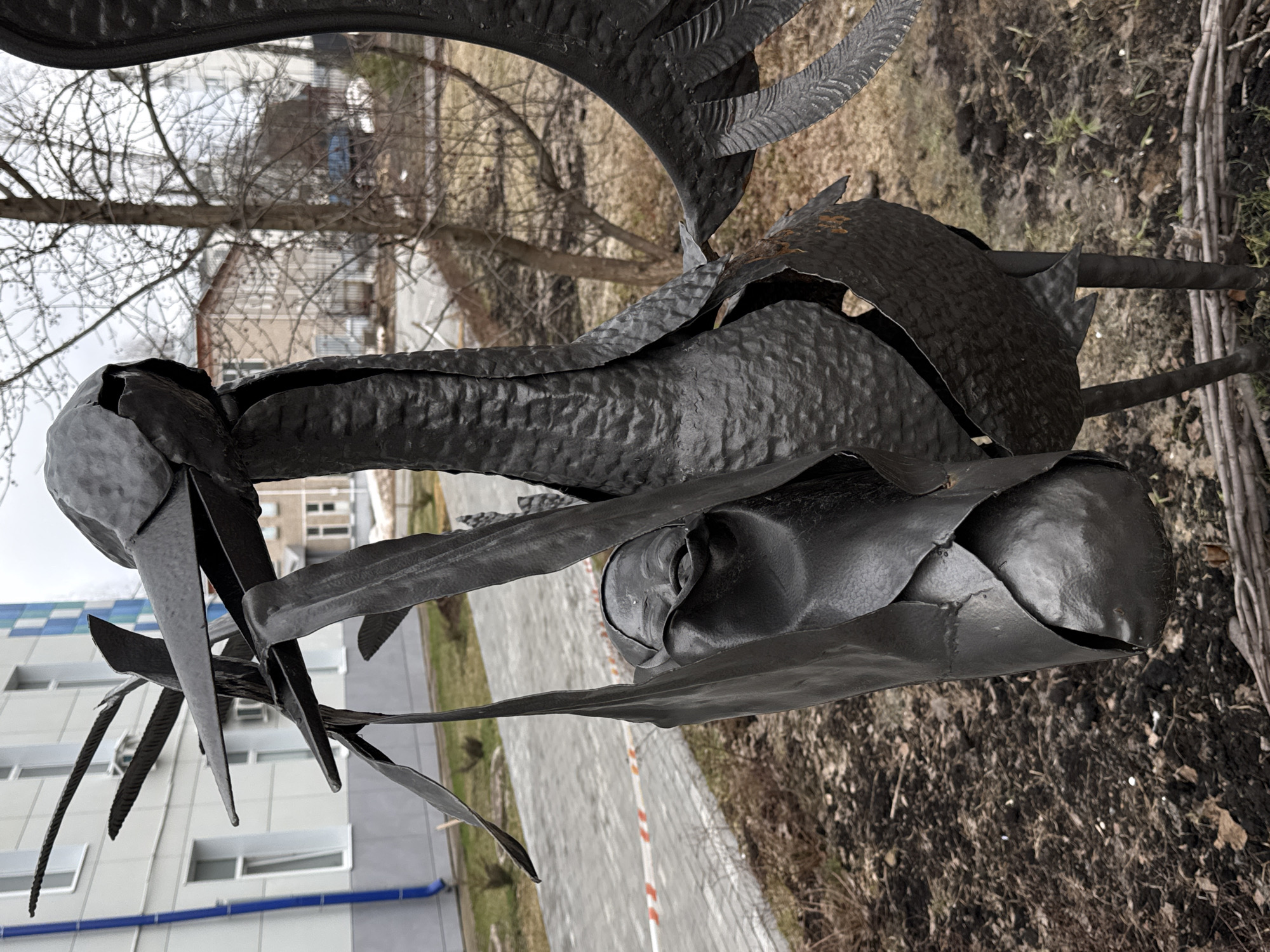 A metal sculpture of a crane is the main subject, set against a backdrop of an urban environment. The sculpture is black, appears to be made of hammered metal, and depicts a crane with intricate details, including textured feathers and a detailed head. The crane is standing near a paved walkway with a red and white striped border. The background includes a multi-story building with a blue and white facade, and bare trees suggesting it is late fall or early spring. The ground around the sculpture is covered with dry grass and leaf litter. The location is in a city, but the specific city and state are not discernible.