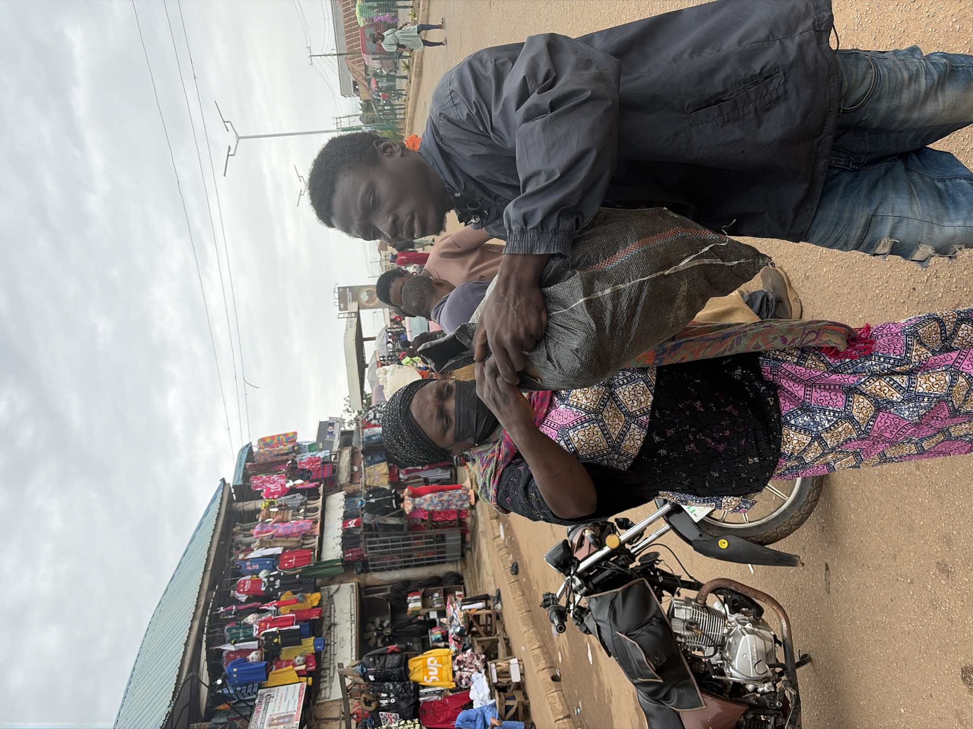 In Bukuru, Nigeria, a market scene unfolds on a dusty street with a stall displaying an array of colorful clothing. Two individuals, a woman wearing a black head covering, a patterned dress, and a face mask, and a young man in a dark jacket and distressed denim jeans, are engaged in an exchange, holding a large, dark-colored plastic bag between them. The woman's hands are occupied with the bag, while the man's right hand is also on it. Behind them, a motorcycle with a brown seat is parked, and other market-goers and stalls are visible in the background under an overcast sky. The overall atmosphere suggests a busy commercial area.