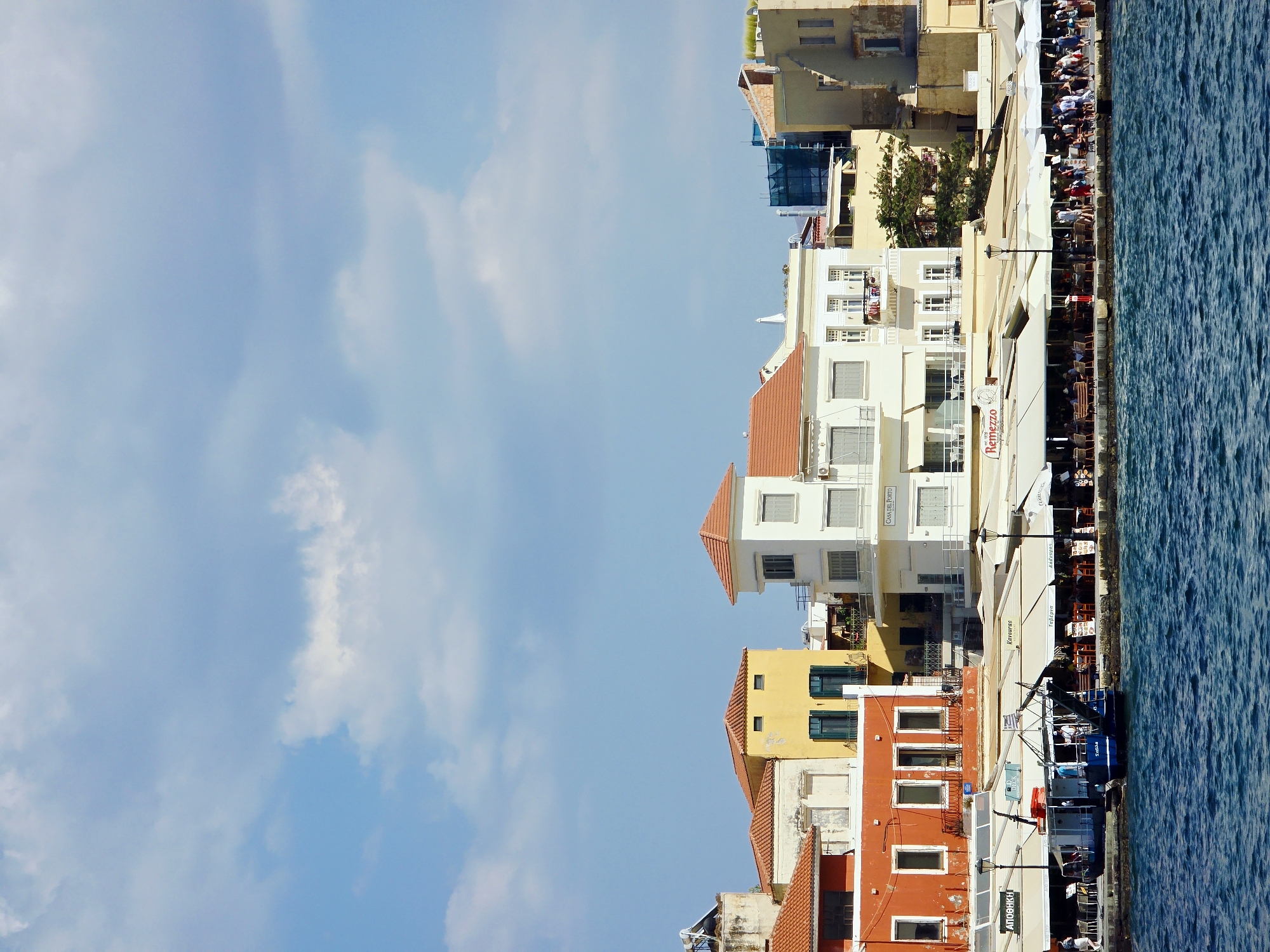This is a vertical, eye-level, medium shot of a waterfront promenade in the city of Chania, Greece, on a bright, sunny day. The foreground is dominated by a large expanse of deep blue, choppy water. A dark blue boat, with "ΣΕΛΙΝΟ" written on its side in white lettering, is docked at the promenade. A crane arm extends from the back of the boat.
The promenade itself features a row of restaurants with outdoor seating, shaded by white umbrellas and awnings. People are seated at tables and walking along the promenade. The buildings lining the promenade are a mix of architectural styles and colors, including white, yellow, and red. Many of these buildings have balconies and open windows. The sky above is a bright blue with scattered white clouds. The scene depicts a lively and picturesque waterfront, likely a popular tourist destination.