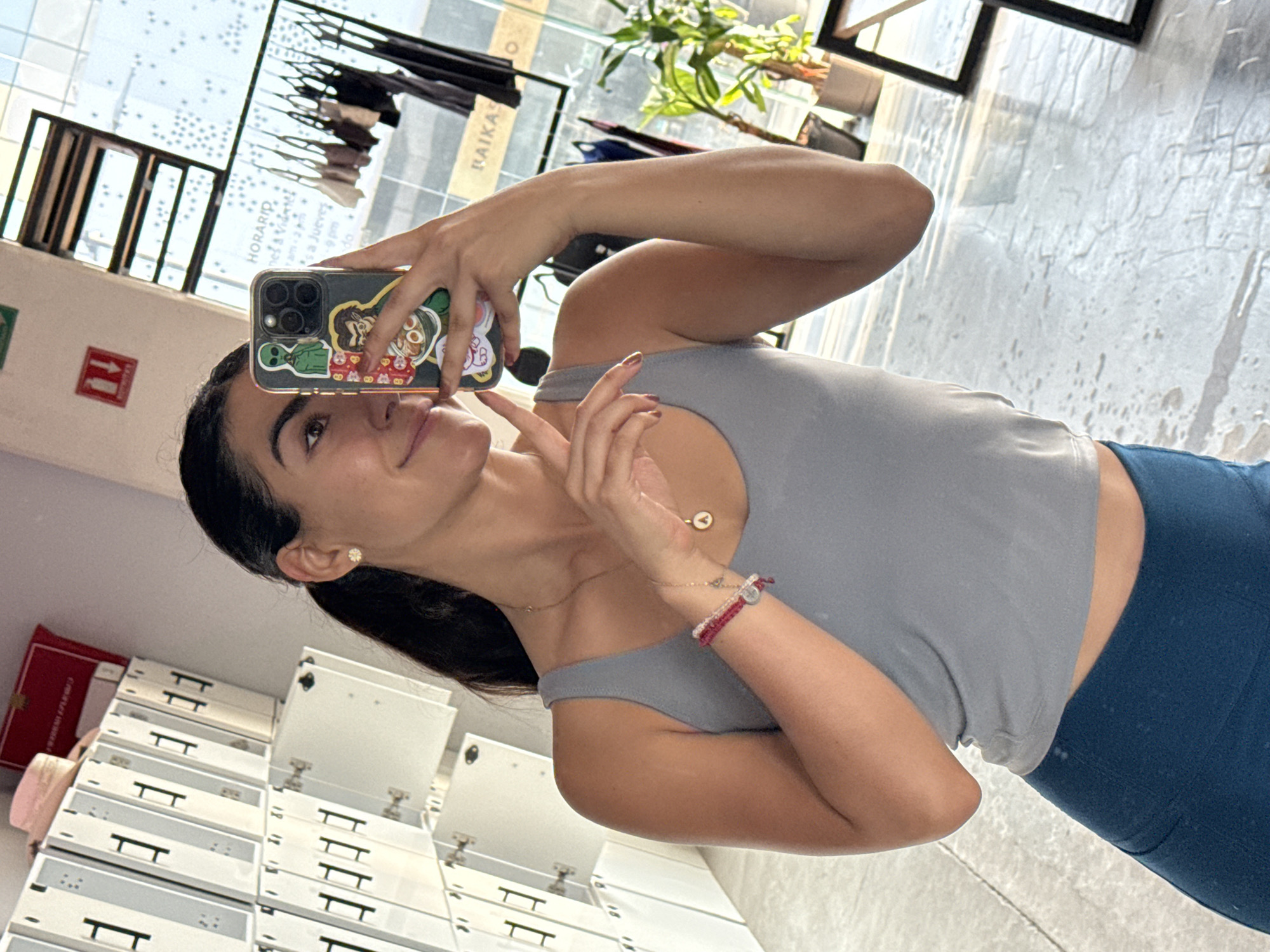 A young woman takes a mirror selfie in an indoor commercial setting, likely a gym or studio in Puebla, Mexico. She is captured from the chest up, looking upward with a subtle smile, her dark hair pulled back. She wears a light grey tank top and dark blue leggings, accessorized with a small stud earring, a delicate necklace with a round pendant, and multiple bracelets on her left wrist, including red beaded ones.
She holds a modern smartphone, enclosed in a clear case adorned with various colorful stickers, including a green alien and cartoon characters. Her left hand is positioned near her chest.
The background, viewed through a large window or glass partition, shows reflections of an indoor space with modern architecture. Visible elements include a black clothing rack with garments and a green potted plant. Text on a reflected sign reads "RAIKA" and "HORARIO" (schedule), with partial details like "Lunes a Viernes" (Monday to Friday) and "a Jueves" (to Thursday), indicating business hours. A red emergency exit sign with an arrow is also reflected. Below, a stack of white and grey modular locker units or drawers are visible. The scene is brightly lit, suggesting it is daytime.