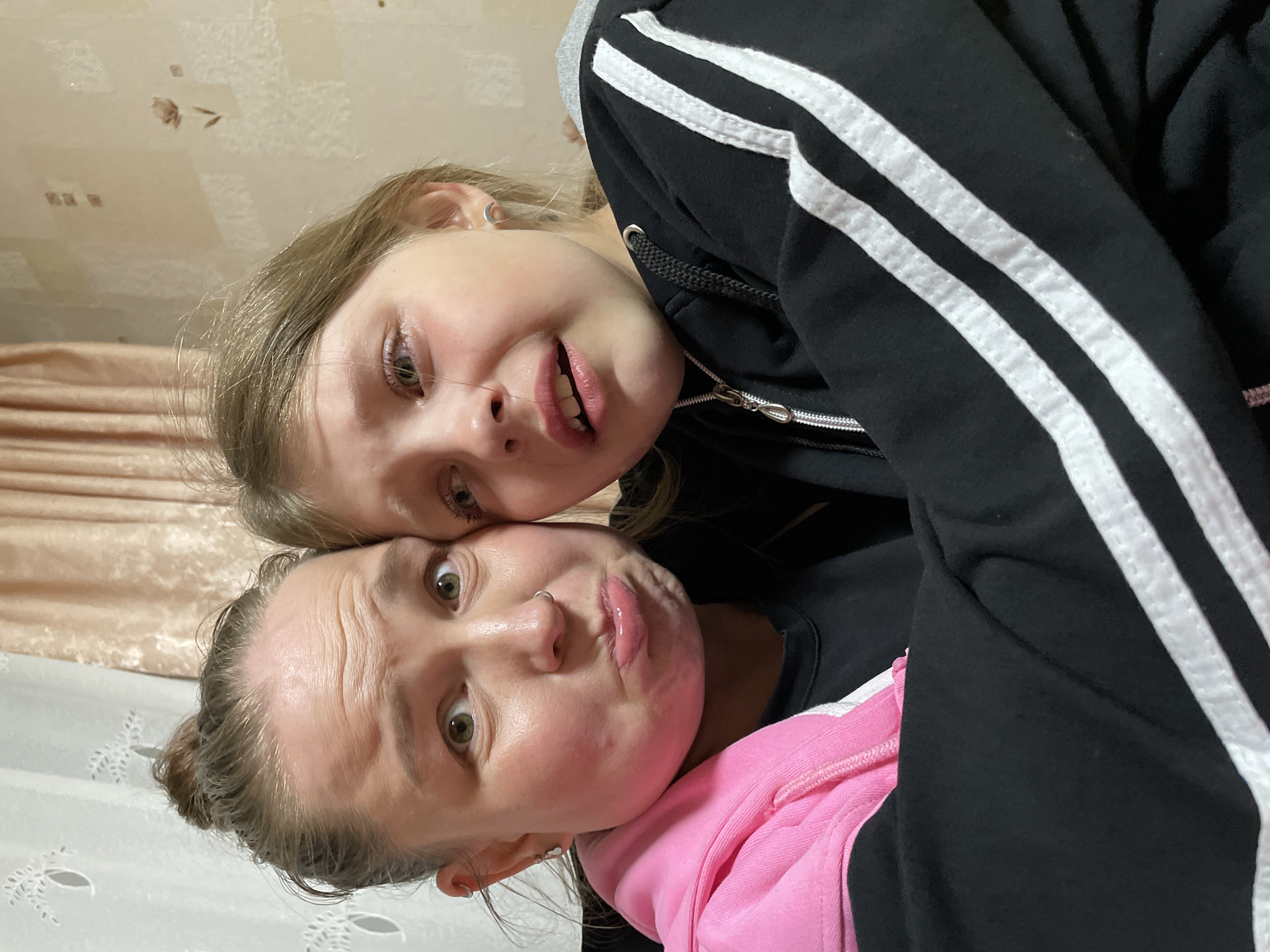 Two individuals, a young girl and an adult female, are close together, possibly taking a selfie. The girl is positioned above, wearing a black hooded sweatshirt with white stripes. The adult female is below, wearing a pink hooded sweatshirt, her hair pulled back. Both faces are visible, displaying exaggerated expressions. The background features a light-colored wall with a decorative pattern. The scene appears to be indoors. There is no information to indicate that the image was taken in Izhevsk, Russia.