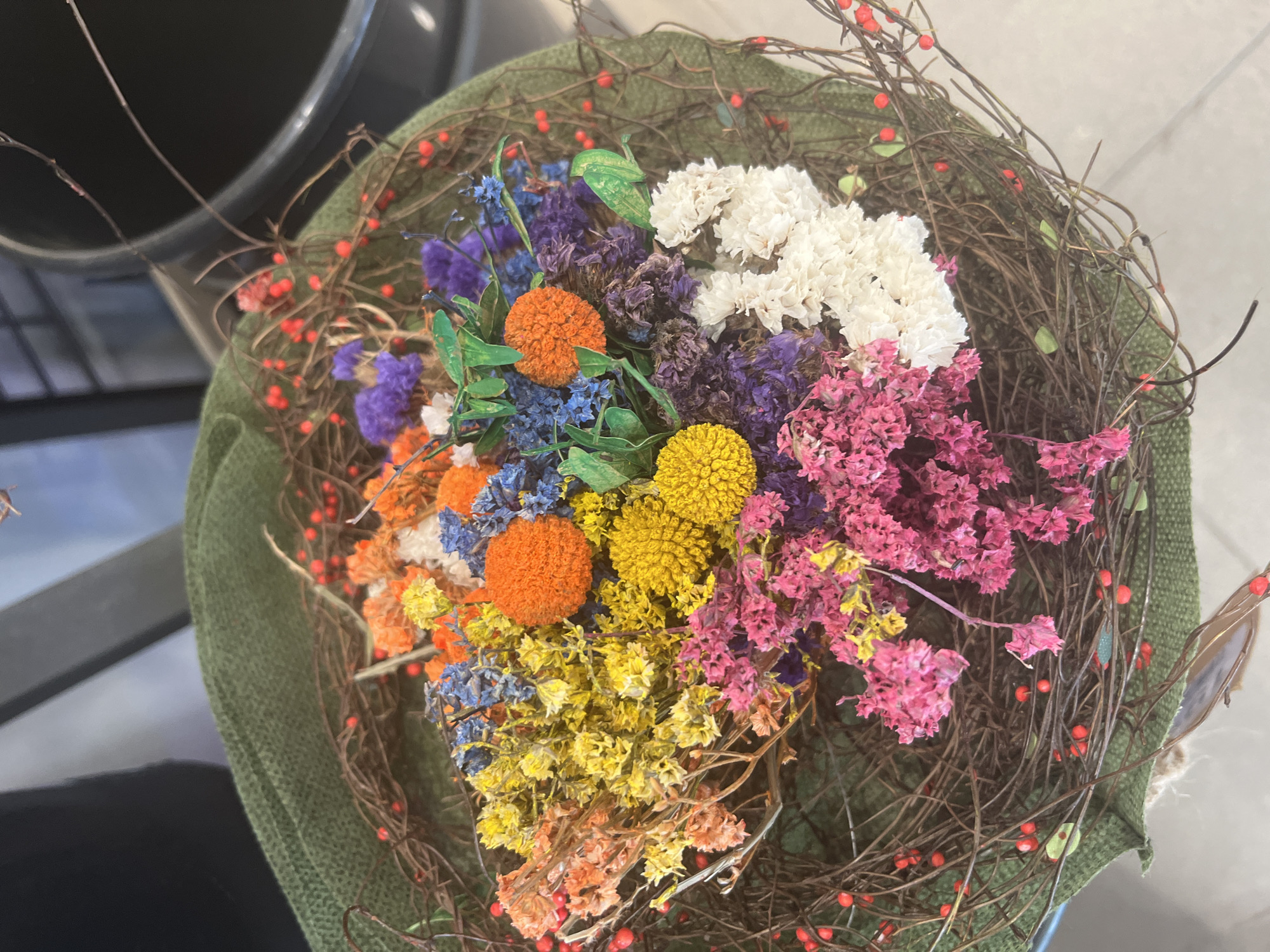 The image shows a bouquet of dried flowers in a round container lined with green fabric. The flowers are a variety of colors including white, pink, purple, blue, orange, and yellow. There are also small red berries attached to thin brown branches that surround the bouquet. Part of a black trashcan and a table are visible in the background. The setting may be in Urbanització El Cim Del Sol, Spain.
