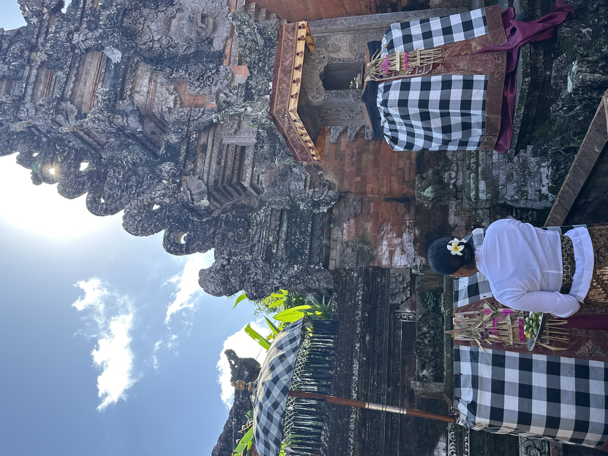 In Ubud District, Indonesia, a woman in a white shirt and patterned sarong stands facing away from the camera, holding a silver tray with offerings. She has a white flower tucked behind her ear. In front of her, a structure is draped in a black and white checkered cloth, adorned with a red and gold textile and a decorative banner. Behind the woman, a large, ornate stone temple structure with intricate carvings rises towards a bright, sunny sky with scattered clouds. To the left, a black and white checkered umbrella stands on a wooden pole, partially obscuring lush green foliage. The scene appears to be a place of worship or cultural significance.