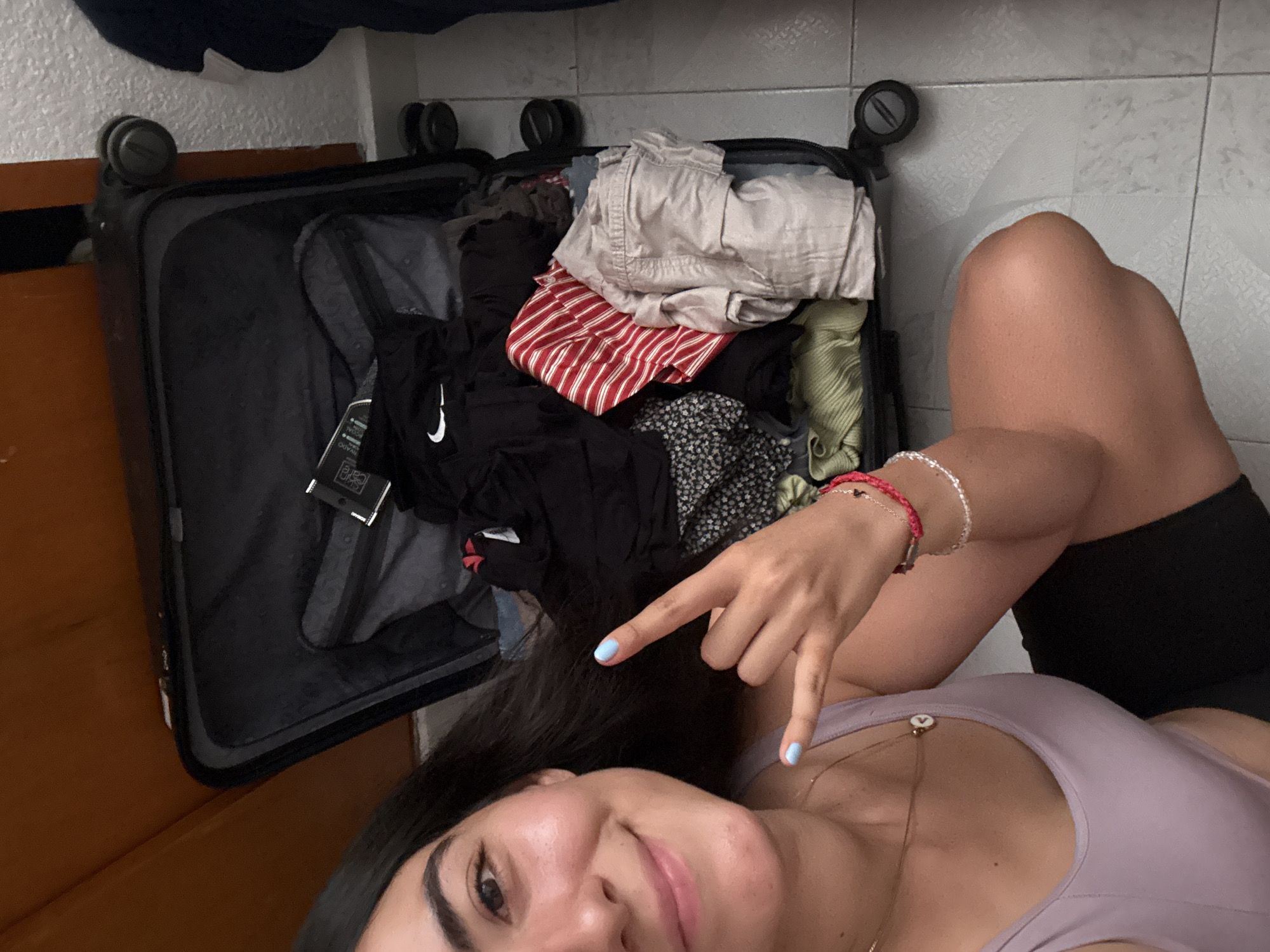 A female lies next to an open, black suitcase filled with clothes. The clothes include a red and white striped shirt, tan shorts, a black Nike brand shirt, and various other garments. The female wears a lilac top, black shorts, and two bracelets on her left wrist. Her fingernails are painted a light blue. The suitcase is located next to a wooden door and a tiled wall. The location is the city of Puebla, Mexico.