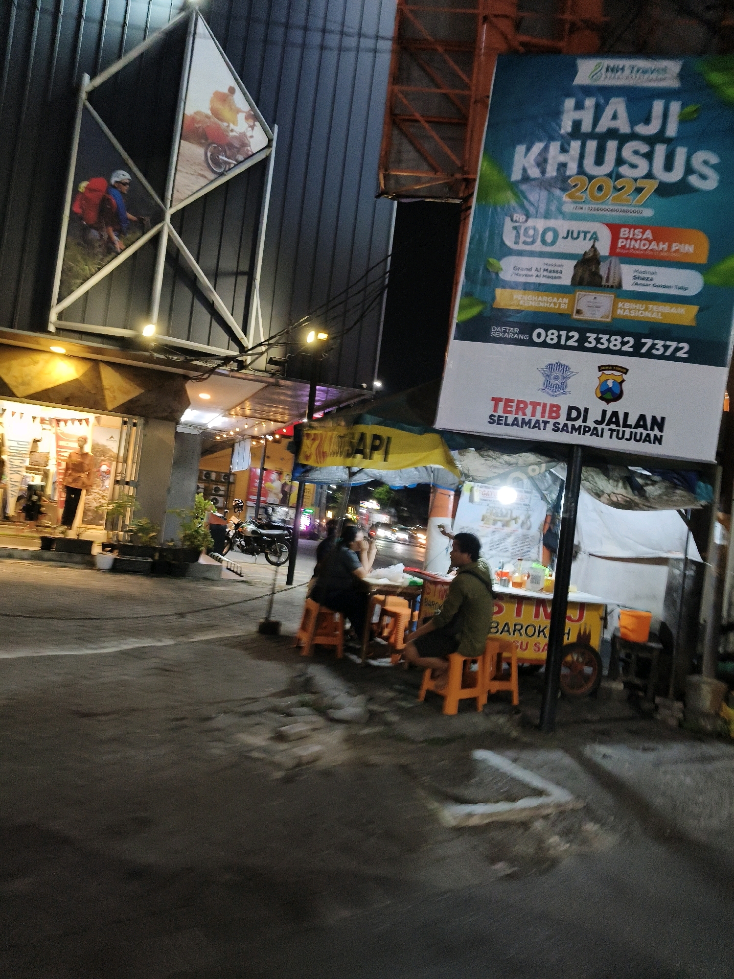 The image depicts a street scene at night, likely in Surabaya, Indonesia, as indicated by the presence of a "Jatim" (Jawa Timur) police emblem. In the foreground, two individuals are seated at a food stall, one facing away from the camera, wearing a green shirt and dark shorts, and the other partially visible, wearing dark clothing. The food stall has a yellow canopy and appears to be selling food or drinks. Next to the stall, an orange plastic stool is visible. Further to the left, a motorcycle is parked.
Dominating the background is a large, orange banner advertising "HAJI KHUSUS 2027" (Special Hajj 2027), offering packages starting at Rp 190 Juta (190 million Rupiah). The banner also includes contact information for inquiries and registration. Above the banner, the facade of a building with a dark, corrugated metal exterior is visible, with a large geometric frame holding pictures of people engaged in outdoor activities, possibly cycling or hiking. The building's lower section has a storefront with illuminated displays and mannequins. Streetlights and signs illuminate the scene, casting shadows and creating a somewhat gritty urban ambiance. The ground is paved and appears wet in places, possibly from recent rain.